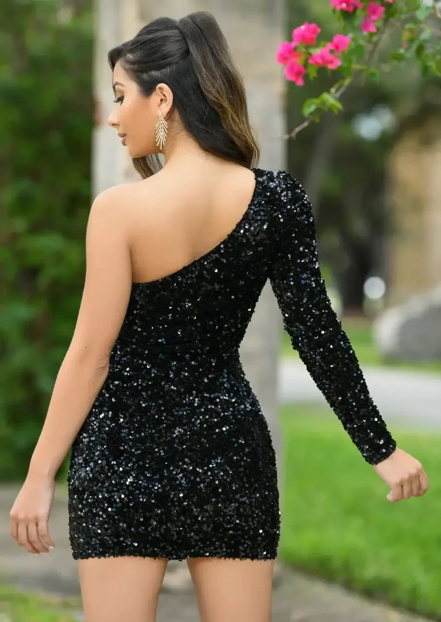 A woman stands with her back turned, wearing a form-fitting black sequined dress with one shoulder exposed. She is outdoors, with greenery and flowers in the background.