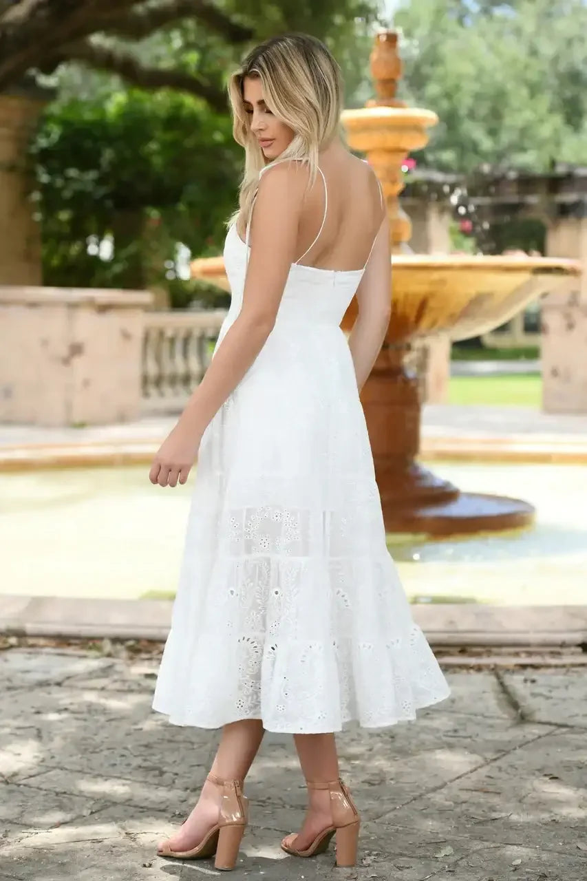 A woman in a white sundress stands with her back to a fountain in a garden, showcasing her flowing dress and strappy heels amidst greenery and soft sunlight.