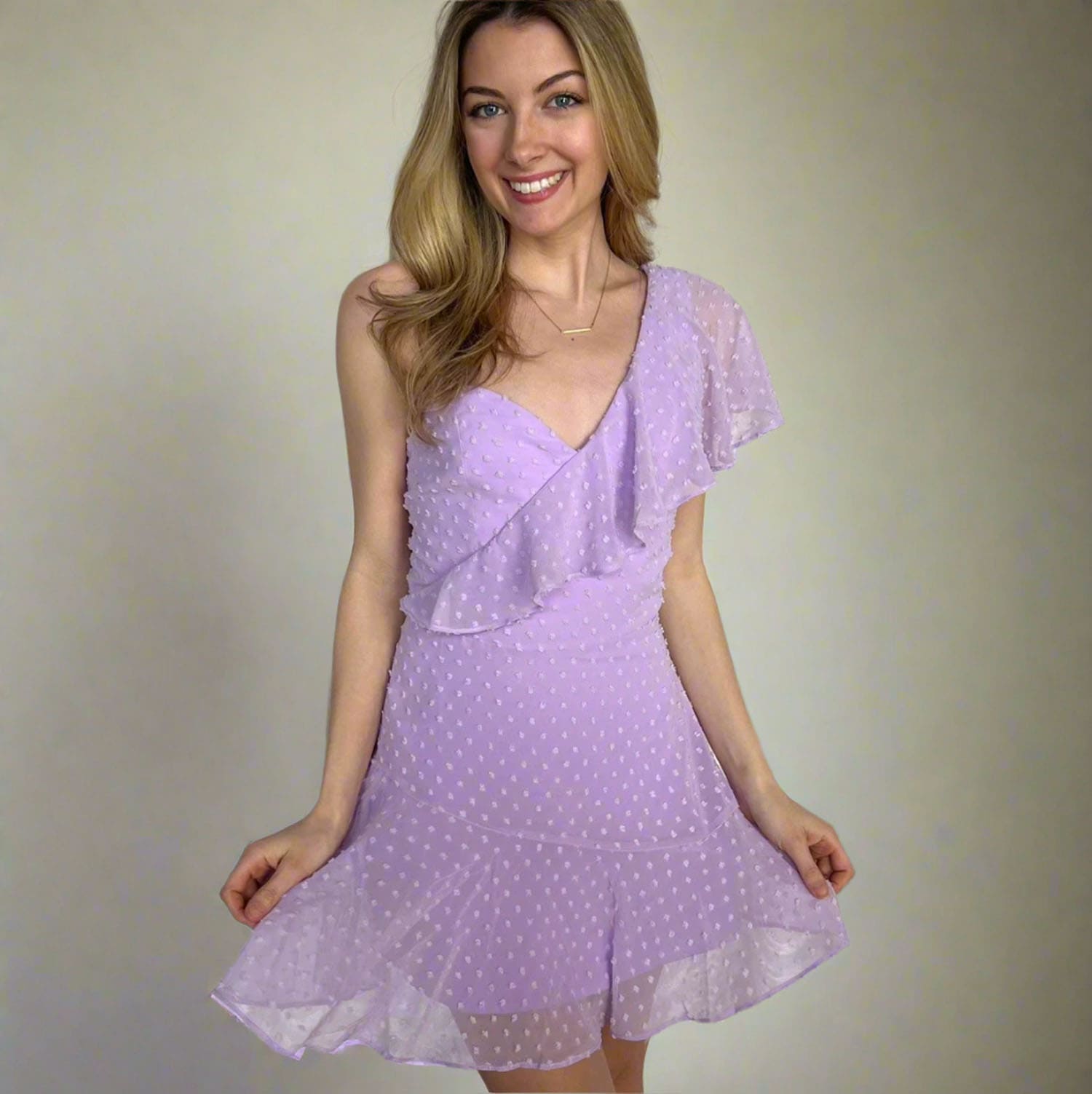 A woman modeling a light purple sleeveless dress with white polka dots, featuring a ruffled wrap-style bodice and flutter sleeves, set against a plain beige studio background.