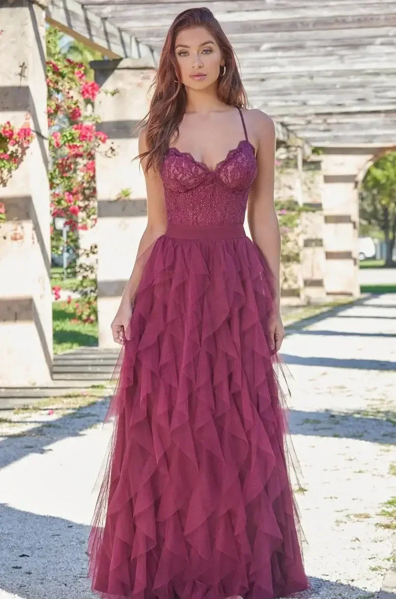 A model stands gracefully in a flowing, burgundy dress adorned with layered ruffles. She poses amidst a floral backdrop and structured columns, under bright sunlight.