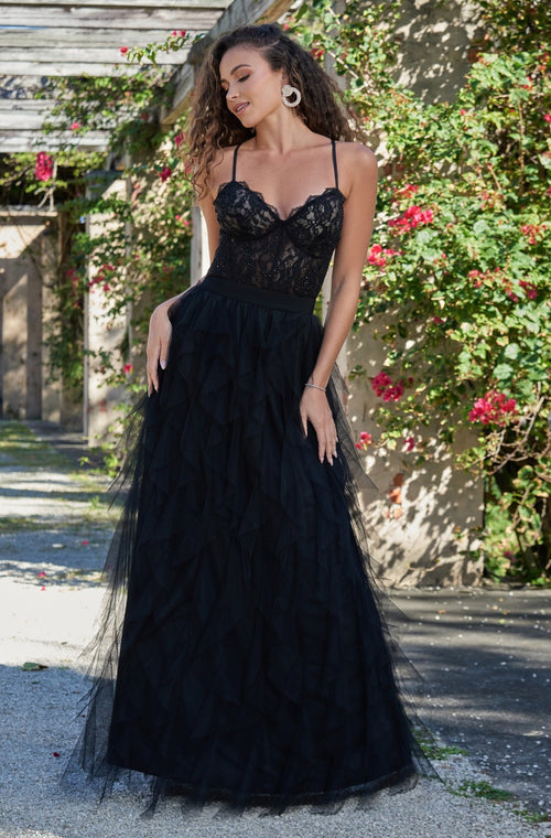 A woman in a black lace and tulle gown stands gracefully outdoors, smiling softly. The environment features flowering vines and rustic architecture, creating a romantic, elegant atmosphere.