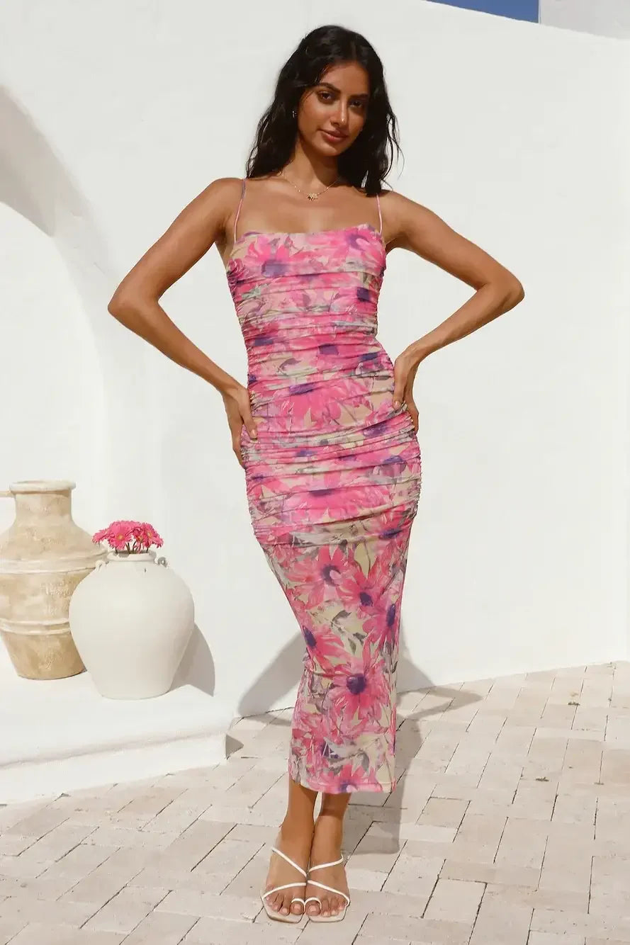 A woman poses confidently in a floral dress with pink hues, hands on hips, against a white wall. Two vases with flowers are visible on the ground nearby.