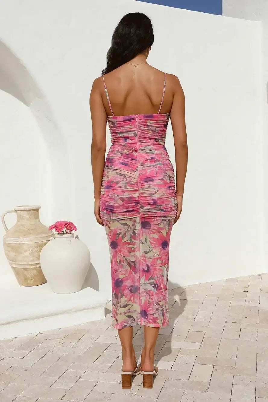A woman stands facing away, wearing a form-fitting, floral-patterned dress with ruching. She is on a sunlit patio, surrounded by white walls and decorative pottery.