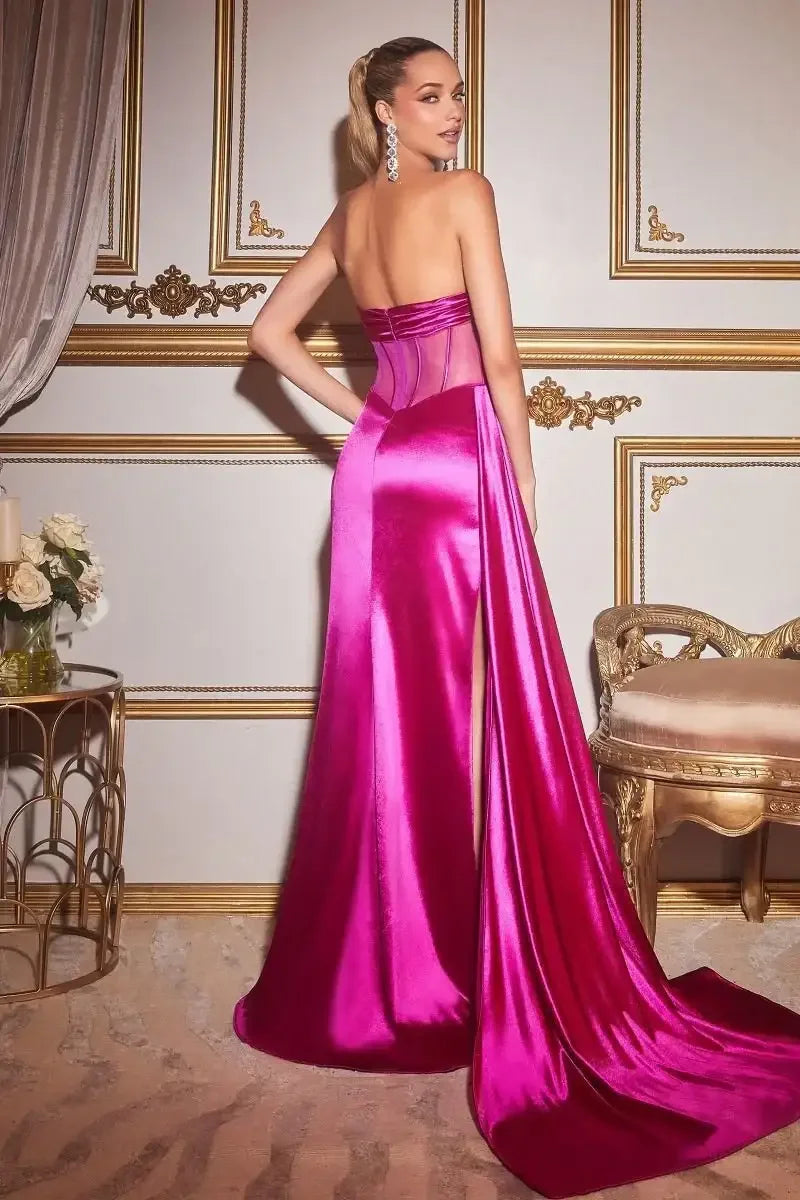 A woman in a striking pink gown poses gracefully, showcasing the dressâ€™s long train. She stands against an ornate, gold-accented wall with elegant decor, including flowers and a gilded chair.