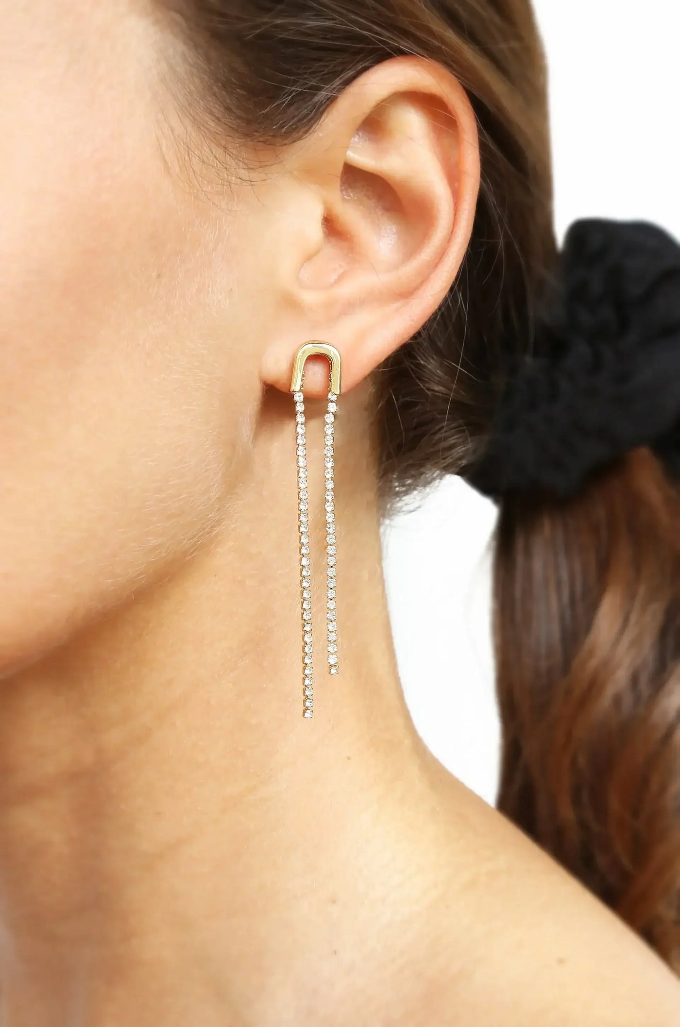 Gold earrings with a U-shaped top and long, dangling chains adorned with sparkling stones are worn on a woman's ear, focusing on her skin tone and hair pulled back.