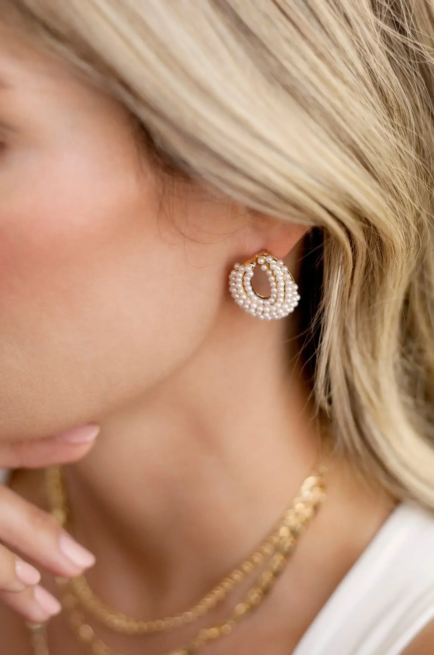 A close-up of a woman's ear features a decorative earring made of pearls and gold. One hand rests thoughtfully against her chin, with soft hair flowing around her face.