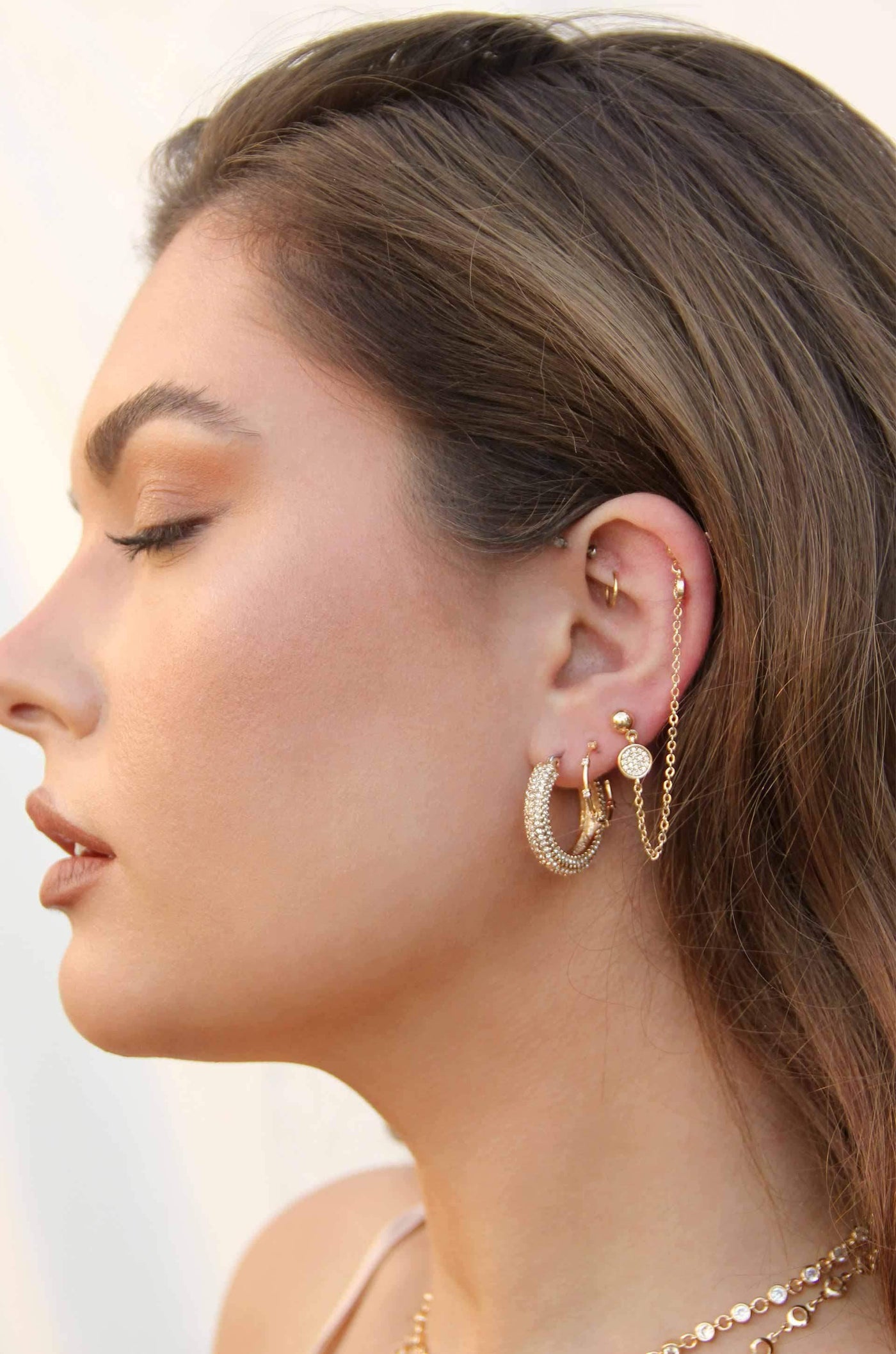 A young woman displays multiple gold earrings in her ear, including hoops and chains, while gazing thoughtfully to the side against a soft, neutral background.