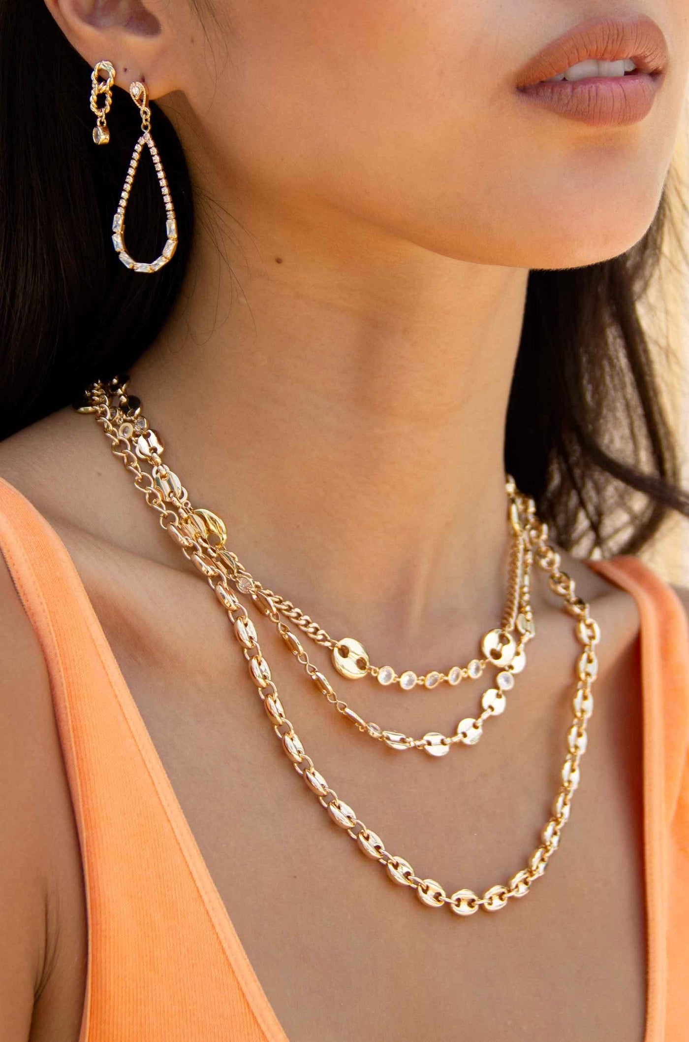 A person wears layered gold necklaces and dangling earrings. They are posed against a light, neutral background while displaying confidence in a casual, orange tank top.