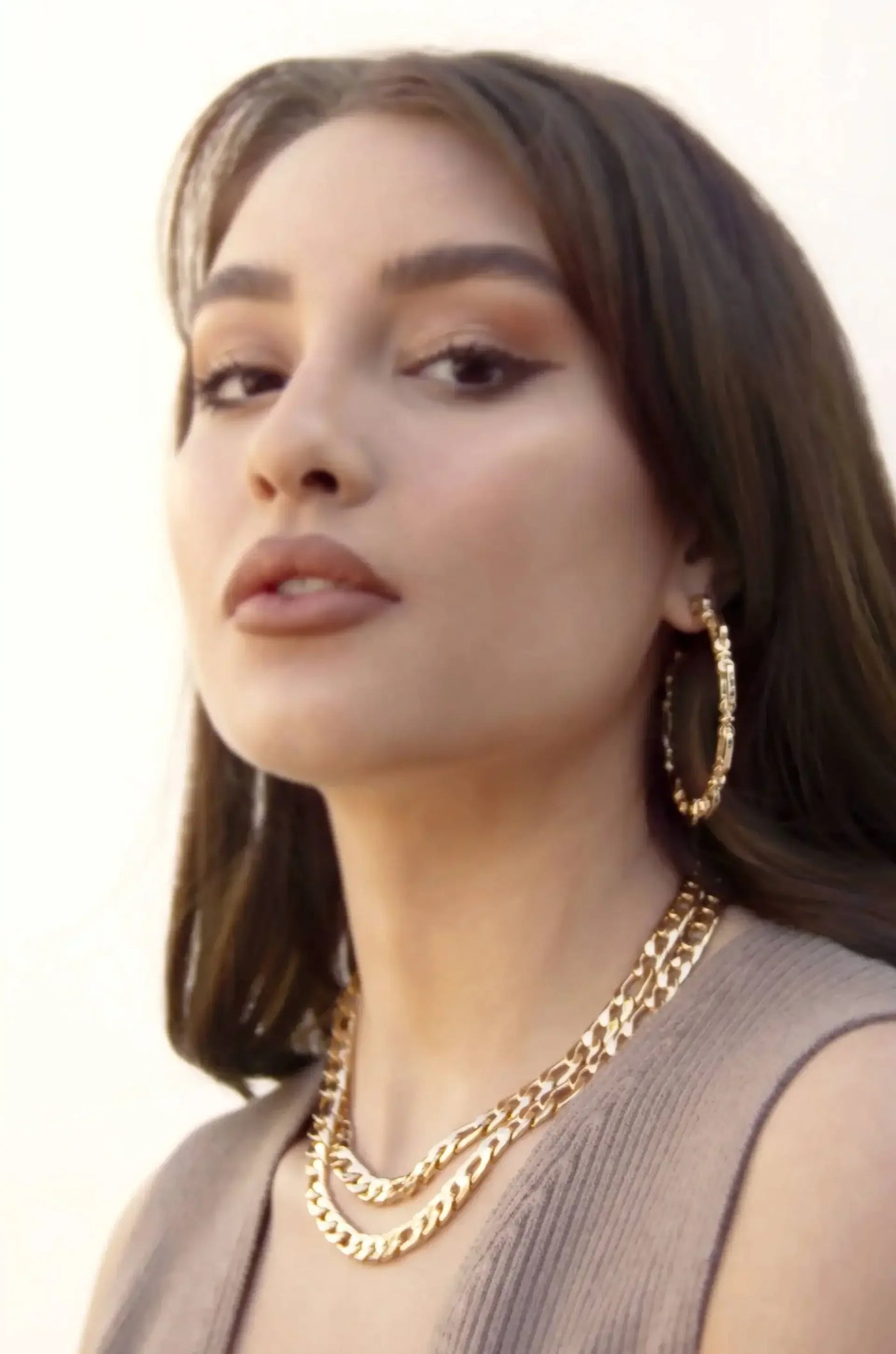 A woman with long, dark hair poses while wearing large gold hoop earrings and layered gold chains. The background is softly lit, creating a warm, elegant atmosphere.