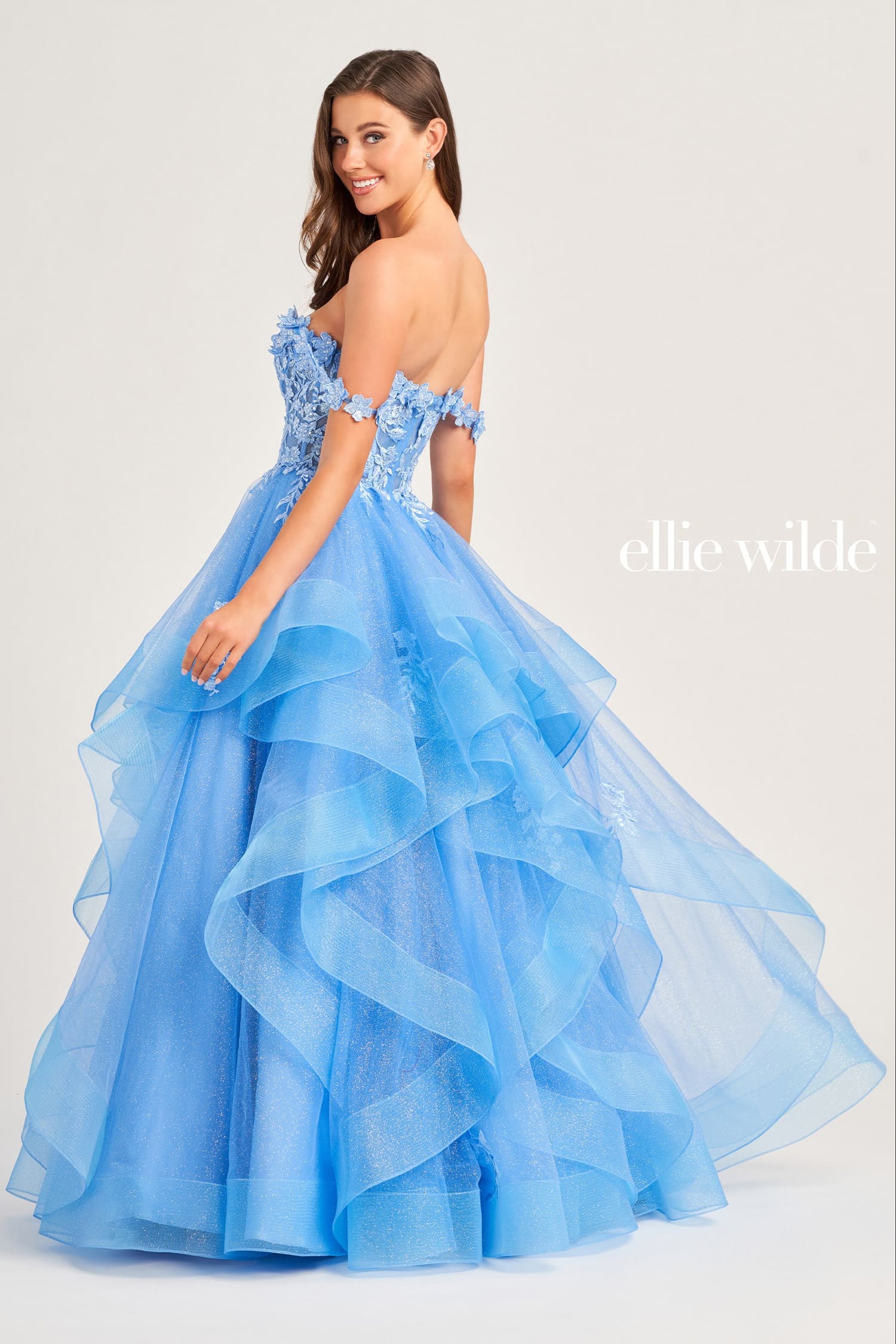 A woman modeling an off-the-shoulder light blue ball gown with a layered ruffled tulle skirt, photographed against a plain light gray studio background. The brand name 'ellie wilde' appears in white text on the right side.