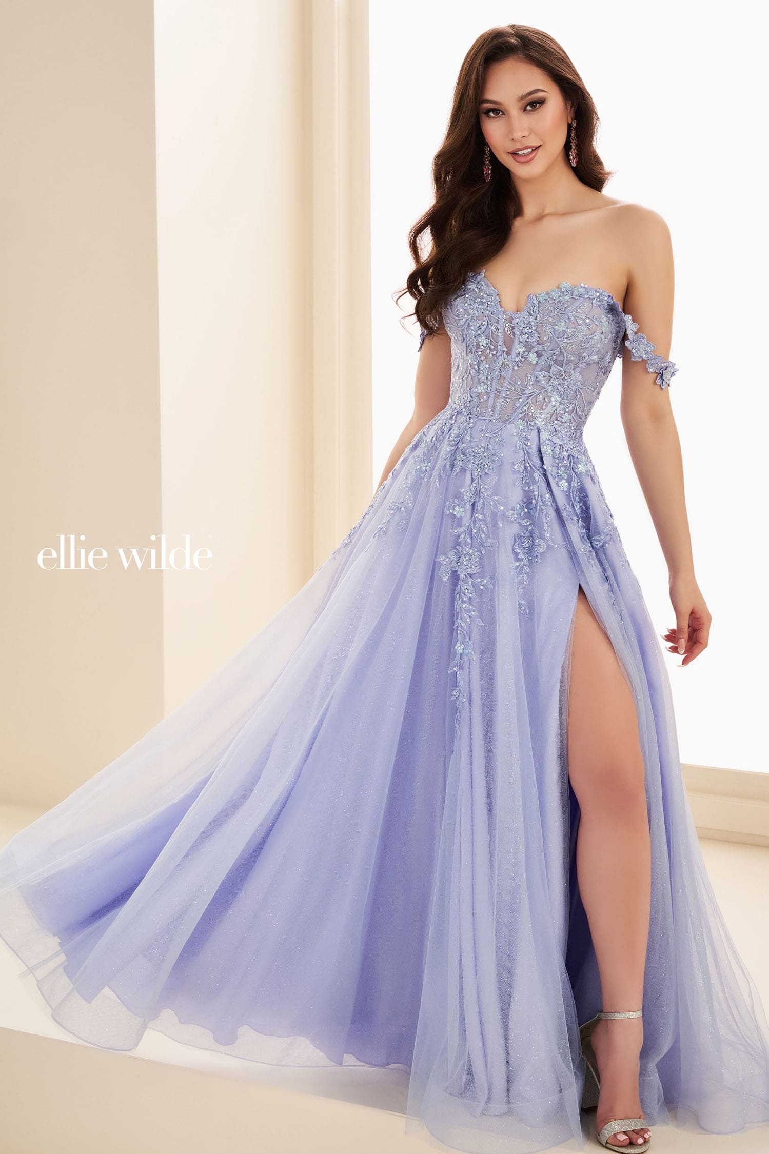 A woman models a strapless light lavender tulle gown with floral lace applique on the bodice and skirt, featuring an off-the-shoulder neckline and a high leg slit. The brand name 'ellie wilde' is printed in white text on the left side.