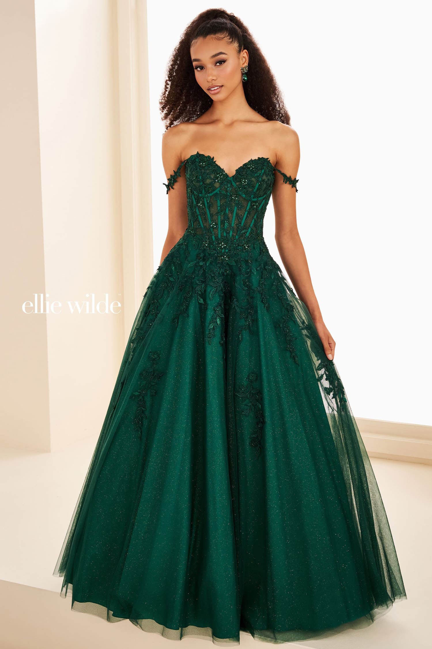 A woman modeling an off-the-shoulder emerald green ball gown with a heavily embellished corset bodice and a full tulle skirt, shown in a brightly lit indoor setting. The brand name 'ellie wilde' is visible as a watermark on the left.