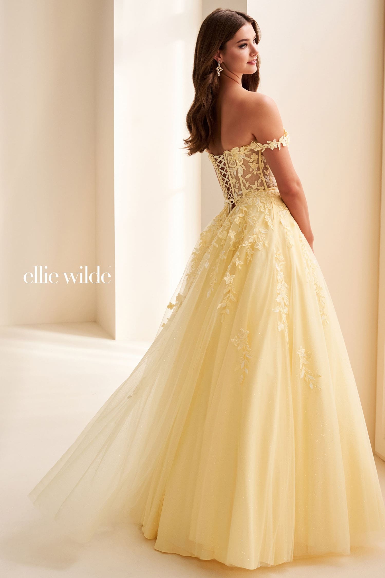 A woman modeling an off-the-shoulder pale yellow ball gown with a lace corset bodice and a full tulle skirt, photographed in a softly lit cream-colored interior. The brand name 'ellie wilde' appears on the left side.