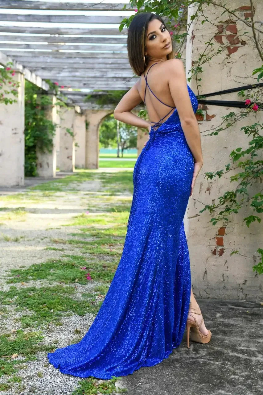 A woman wears a sparkling blue gown and poses confidently, showcasing a scenic outdoor area with arches and greenery in the background, under soft sunlight.