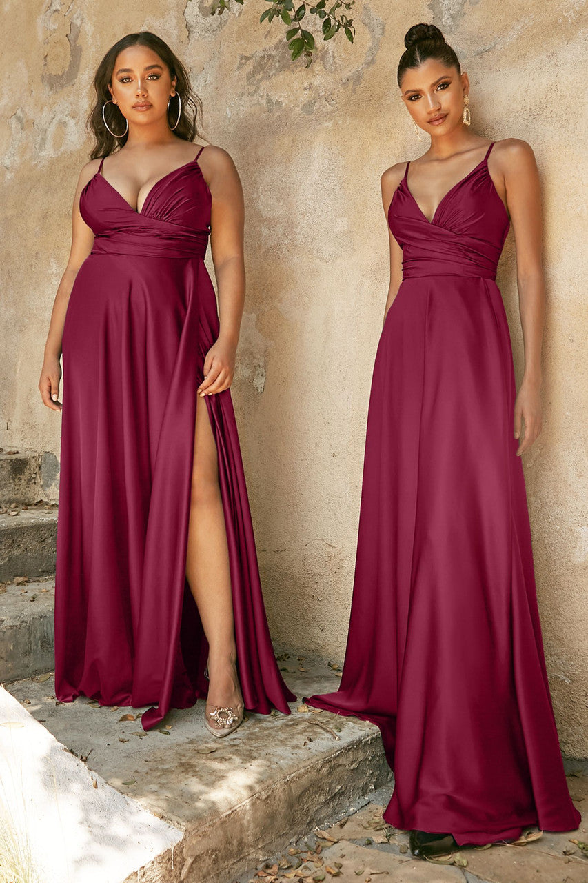 Two women stand side by side on stone steps, wearing elegant burgundy dresses with fitted bodices and flowing skirts. The background features a textured, neutral wall with scattered leaves.
