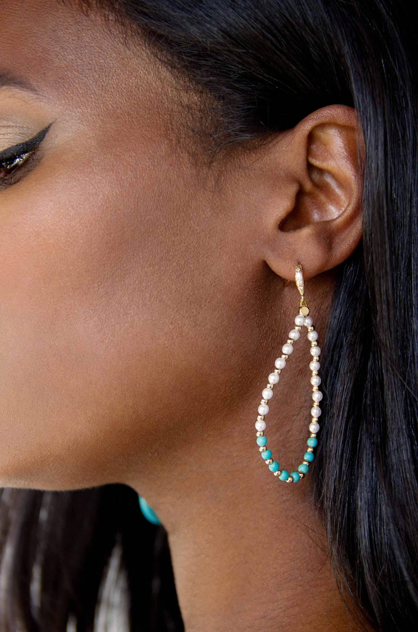 A teardrop earring made of alternating pearls and turquoise beads hangs decoratively from a womanâ€™s ear, highlighting her smooth skin and long black hair against a neutral background.