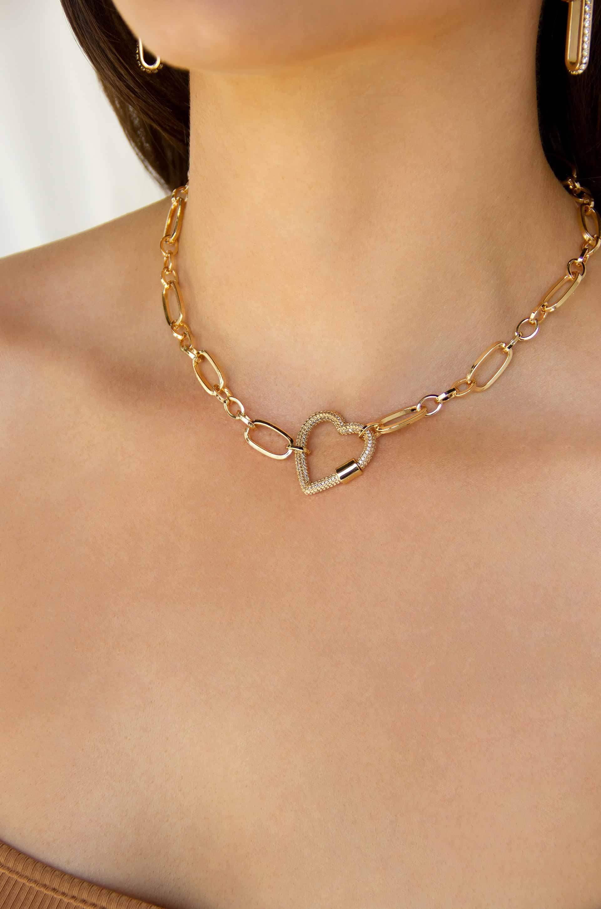 A gold chain necklace with a heart-shaped pendant rests on a bare shoulder, highlighting a smooth skin tone. The background features soft, neutral colors, enhancing the jewelry's elegance.