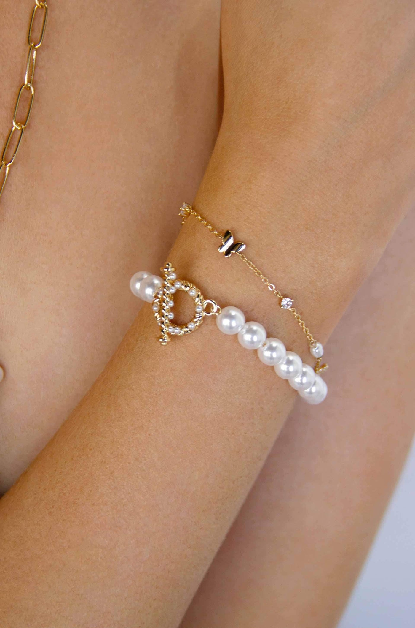 A delicate wrist adorned with two bracelets: one features lustrous pearls and a decorative clasp, while the other is a fine chain with a small butterfly charm, suggesting a stylish, elegant context.