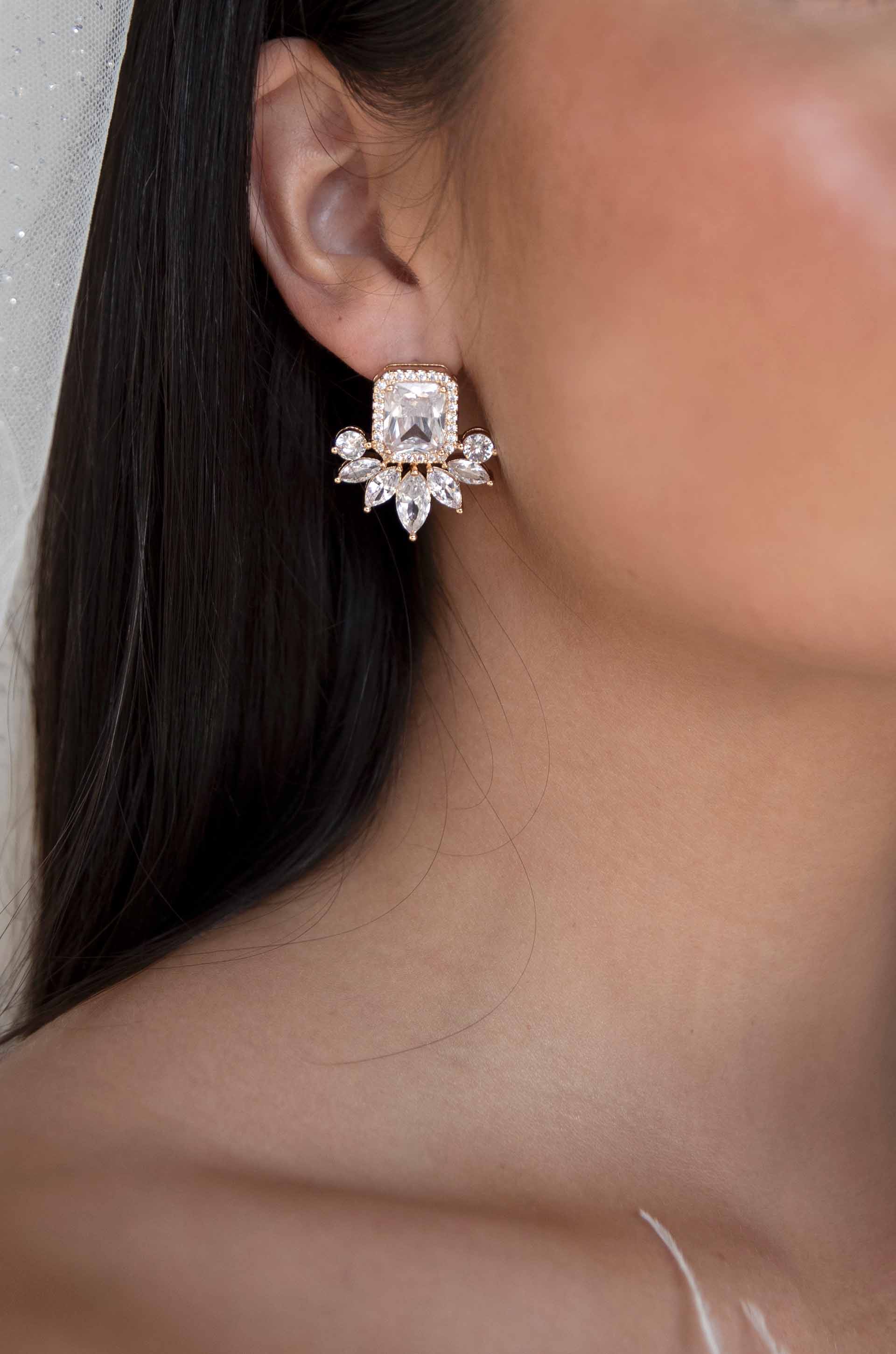 A large, ornate earring is worn on the left ear, featuring a square clear gemstone surrounded by smaller stones. The context includes smooth skin and long, dark hair.