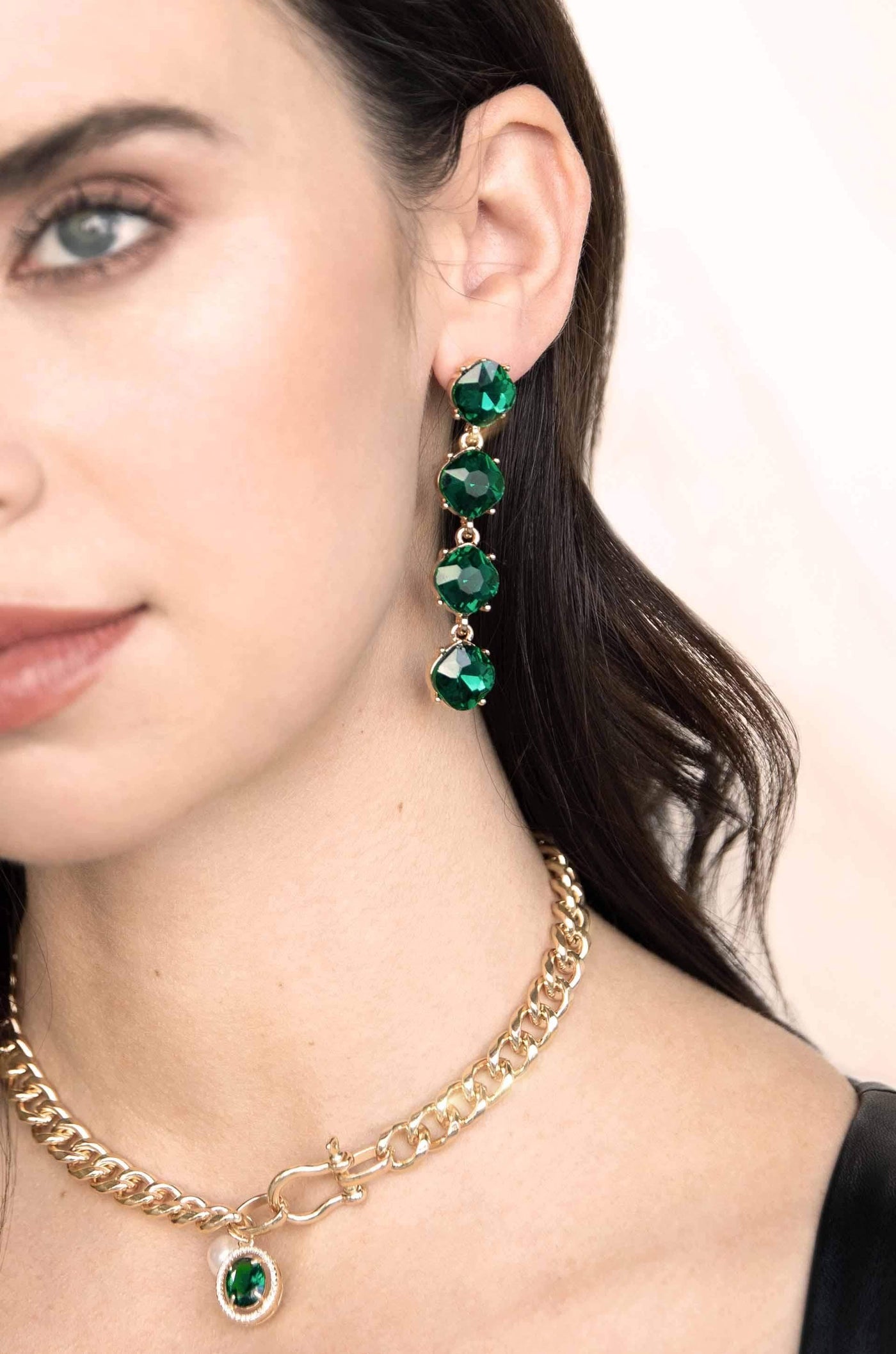 A woman wears a gold chain necklace and a long, emerald-green jeweled earring. The background is light, emphasizing her accessories and features, including her smooth skin and dark hair.