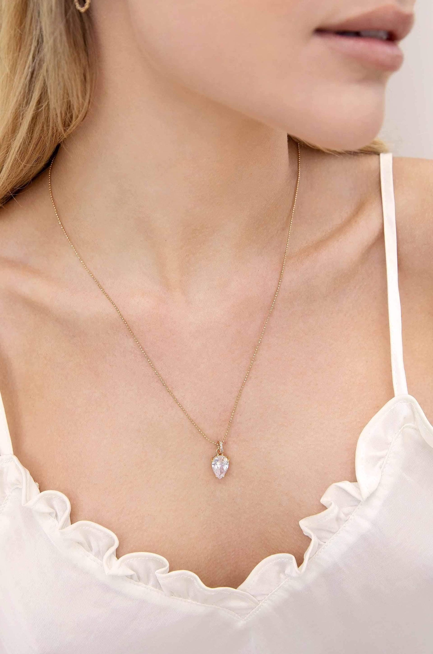 A delicate necklace with a sparkling pendant hangs elegantly around a personâ€™s neck. The individual wears a sleeveless, ruffled white top, with soft lighting accentuating the tranquil setting.
