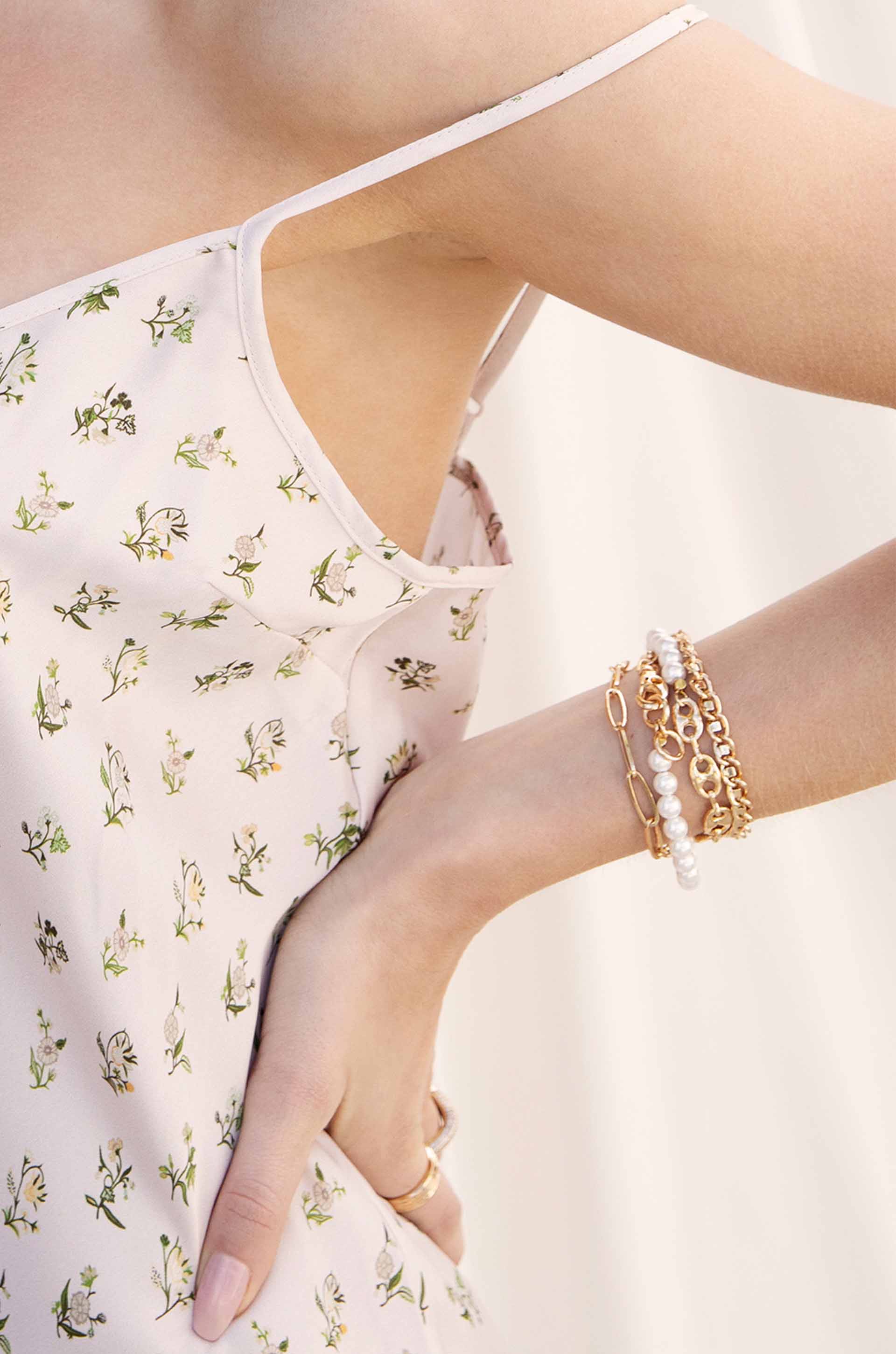 A wrist adorned with multiple bracelets, including gold chains and pearl accents, rests against a floral-patterned fabric, suggesting a casual yet stylish summer setting. The background appears soft and light.