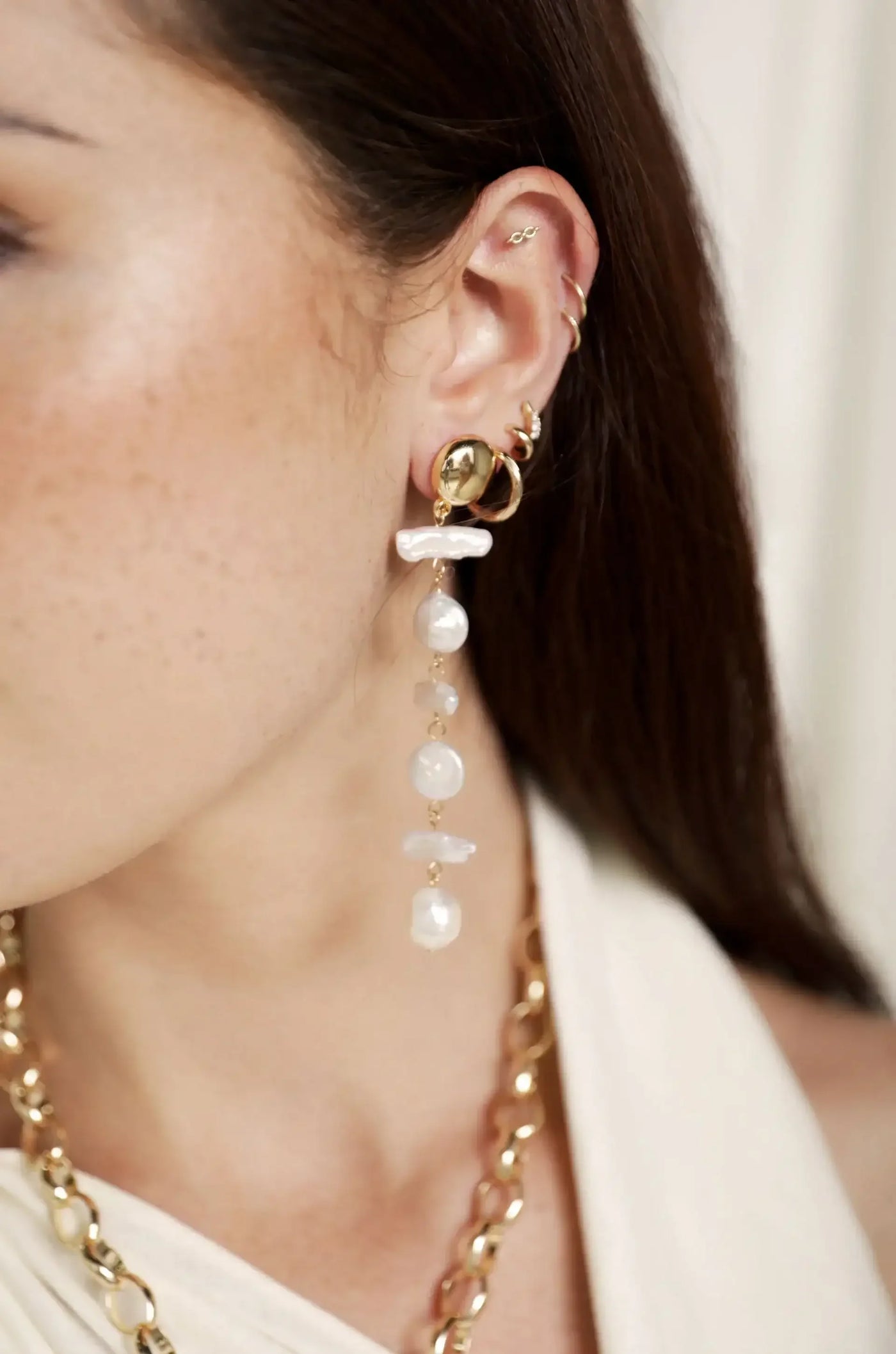 An elegant earring featuring gold elements and dangling pearls adorns a woman's ear. She has multiple ear piercings, and the backdrop is softly blurred, suggesting a neutral environment.