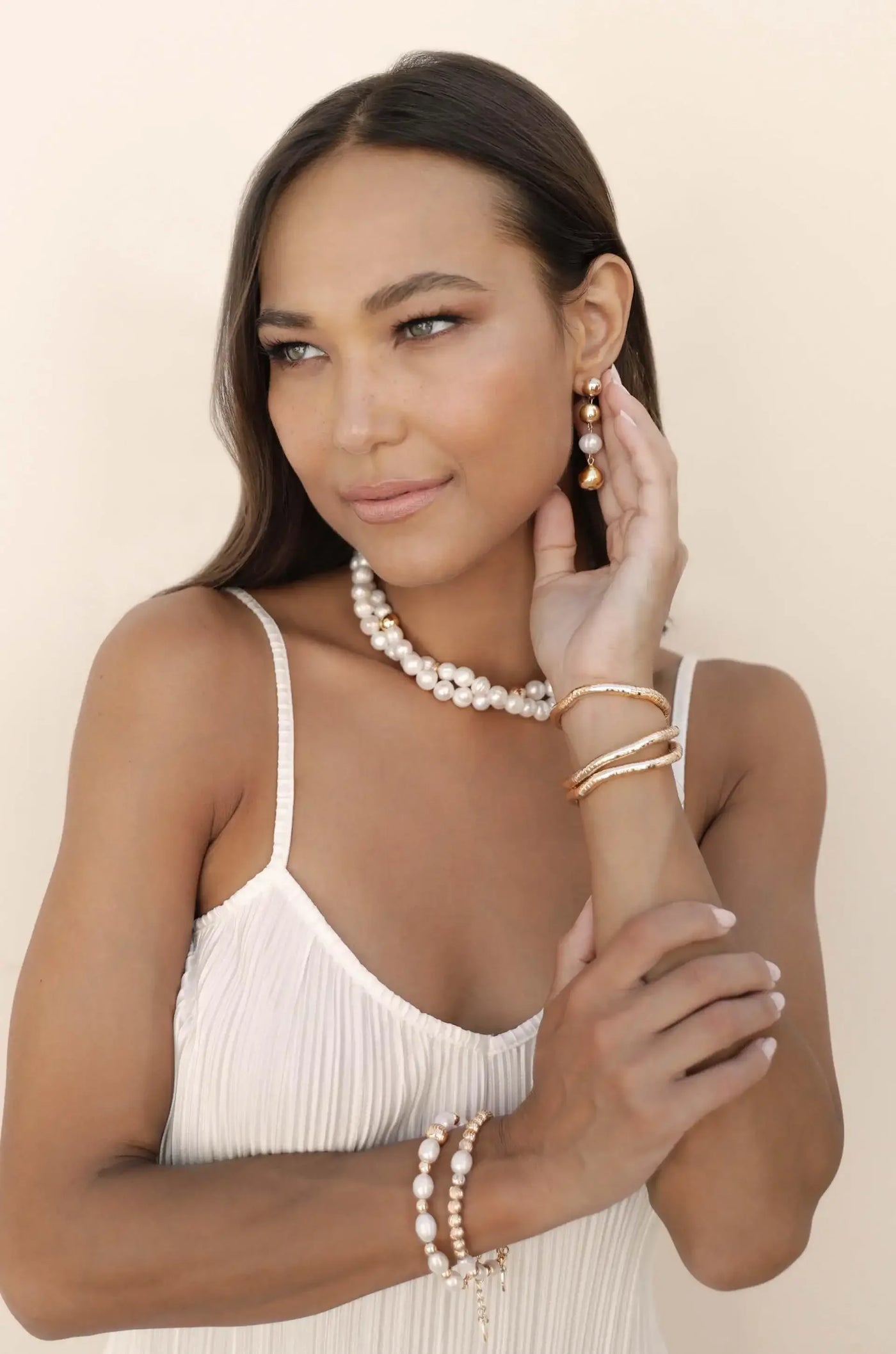 A woman with long hair wears pearl jewelry, including earrings and a necklace. She gently touches her face, set against a soft, neutral background that enhances the jewelry's elegance.