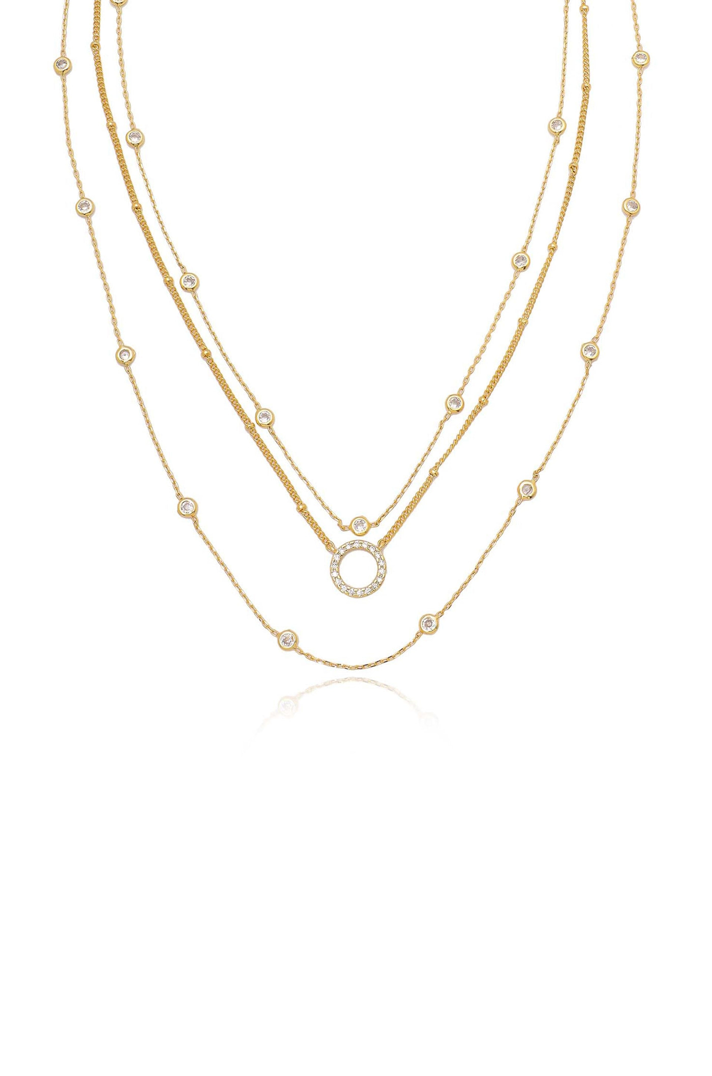 Three delicate gold chains are layered together, featuring round, embellished links and a central circular pendant adorned with stones, set against a plain white background.