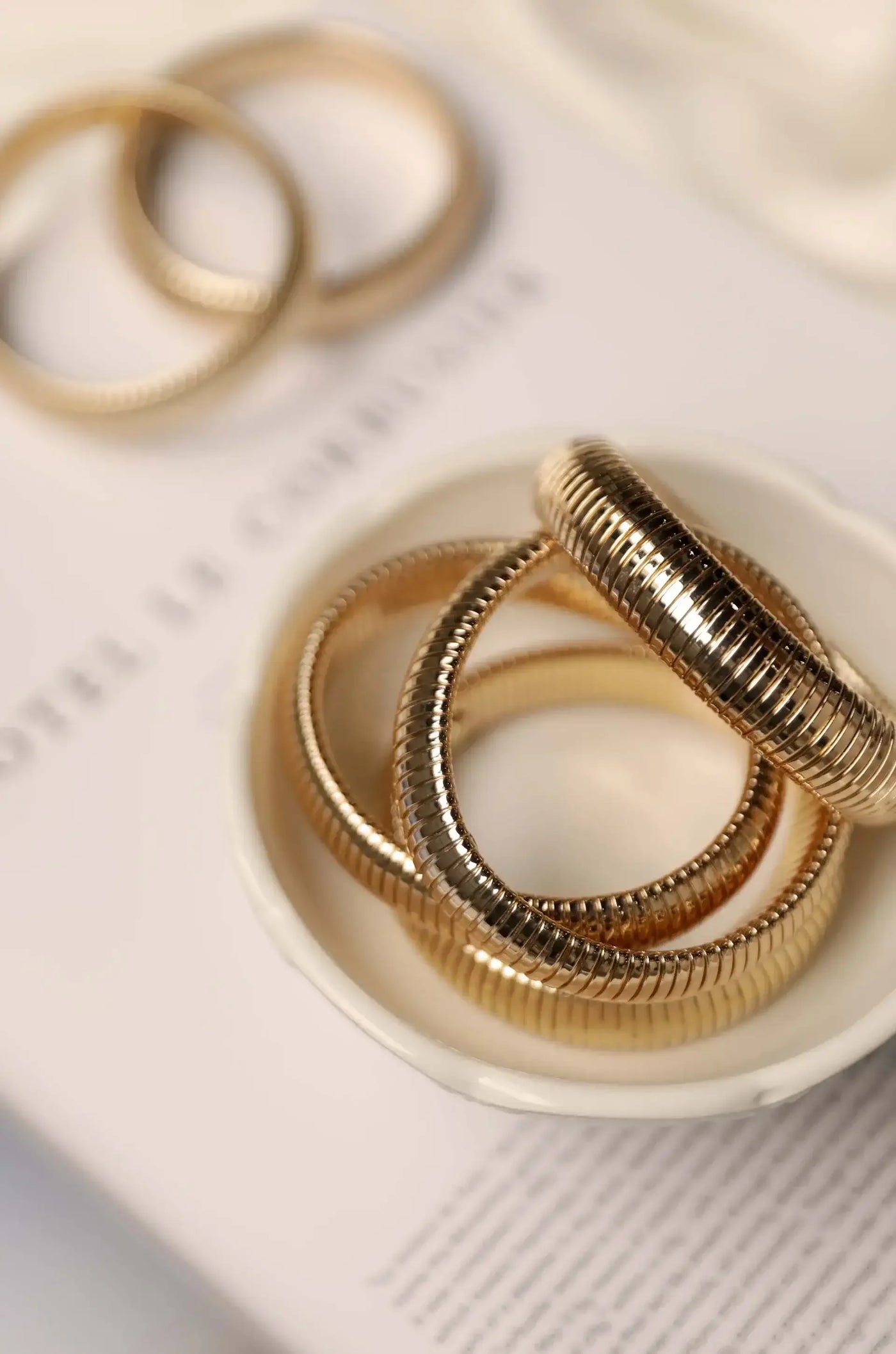 Three metallic bangles are stacked inside a small white dish. They are placed atop a soft, blurred magazine page featuring text, creating a minimalist and elegant display.