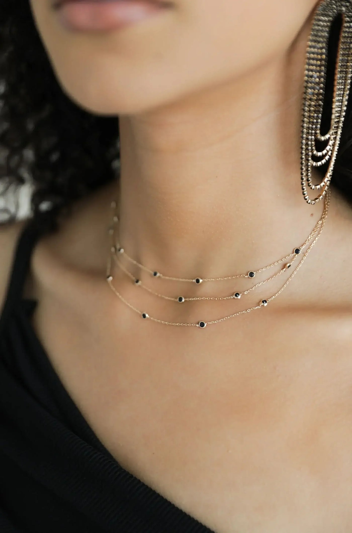 A layered gold necklace with small black beads decorates the neck of a person with curly hair, wearing a dark clothing. The background is softly blurred, highlighting the jewelry.