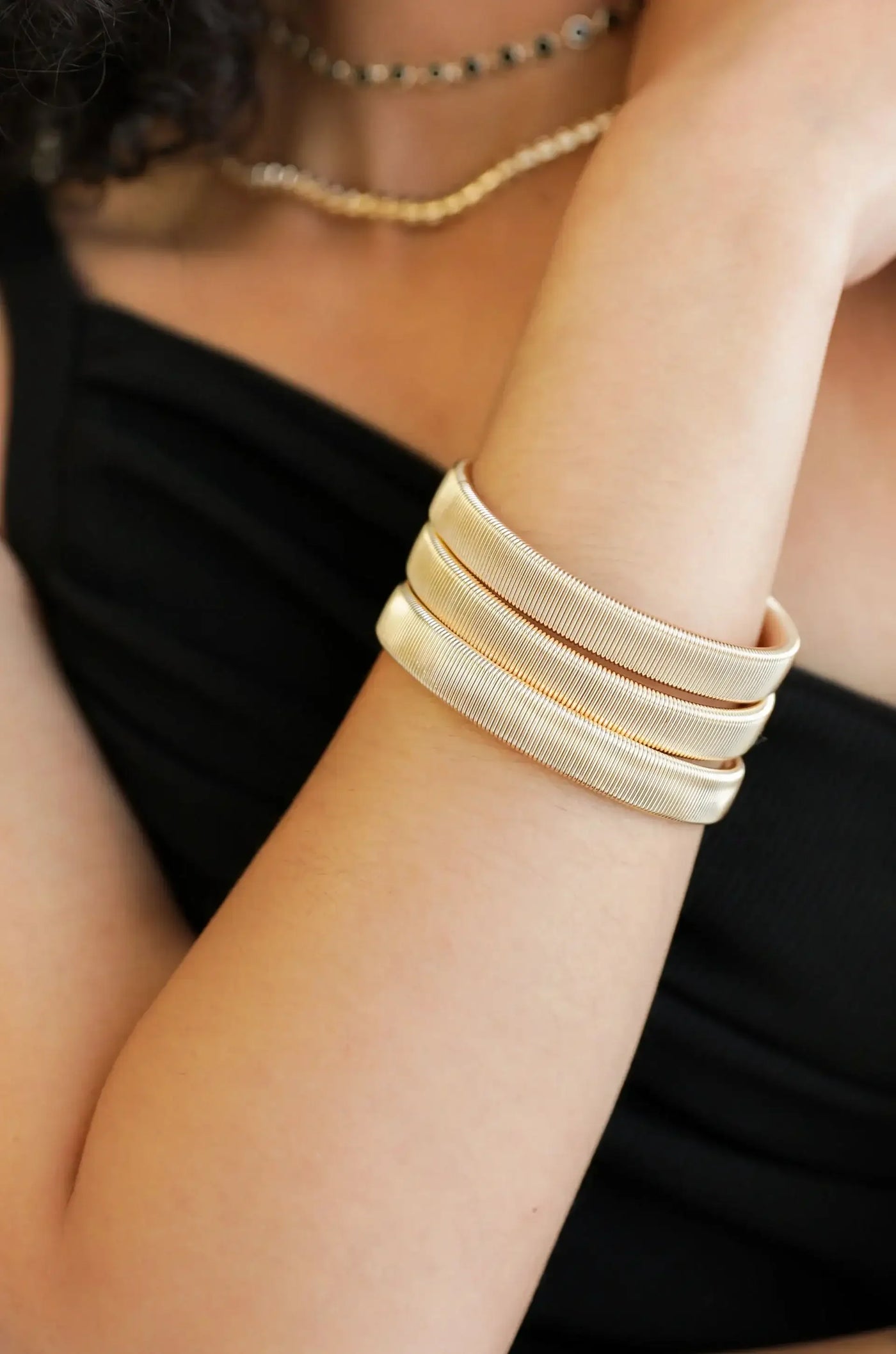 A slender arm adorned with three gold bangles rests against a black top. The bangles are smooth and shiny, catching light, presenting a stylish and elegant look in a close-up view.