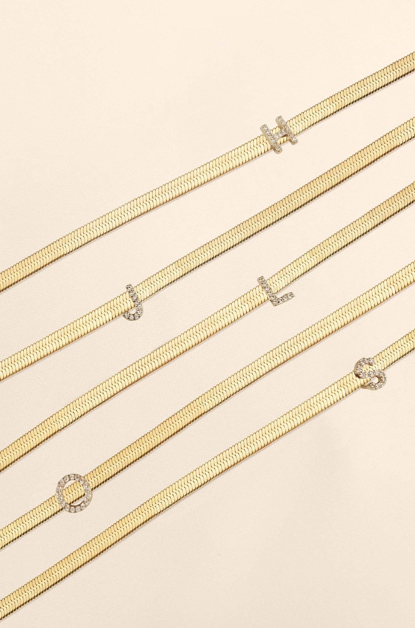 Five gold necklaces with various decorative elements lay flat on a soft, light beige surface. They feature letter-shaped embellishments, creating a personalized design.