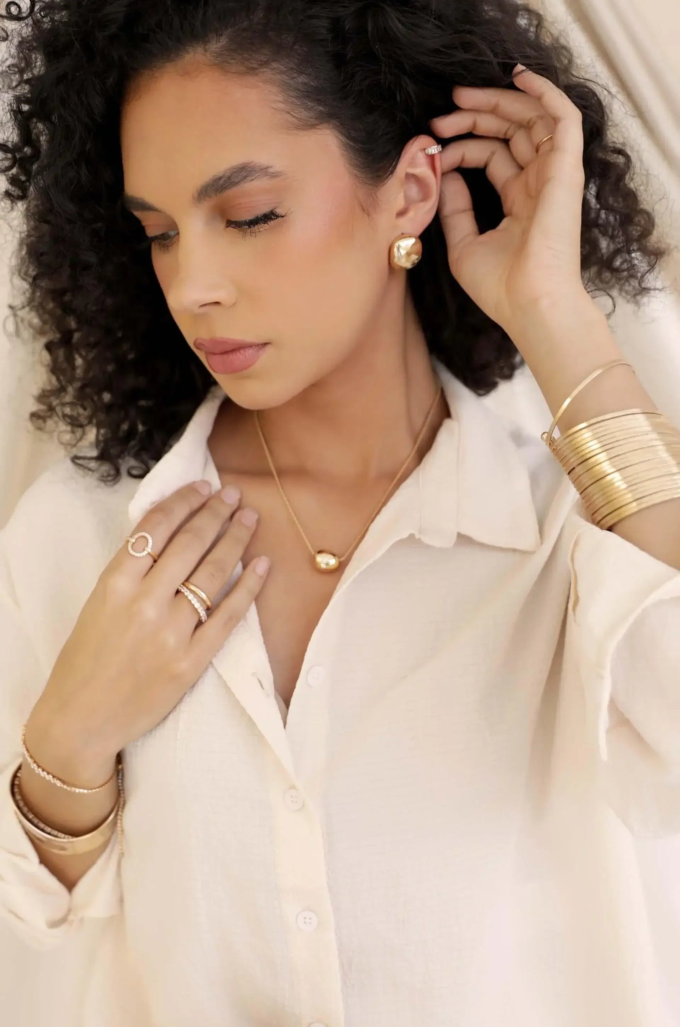 A woman with curly hair wears elegant gold jewelry, gently touching her necklace while dressed in a light, textured shirt. The soft background complements her sophisticated appearance.