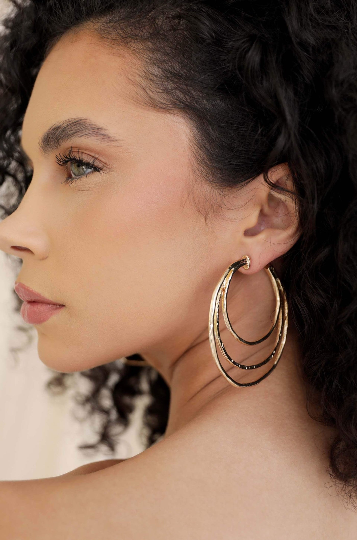 A woman with curly hair wears large, multi-layered gold hoop earrings. She poses sideways, highlighting the earrings against a soft, neutral background that enhances her features.