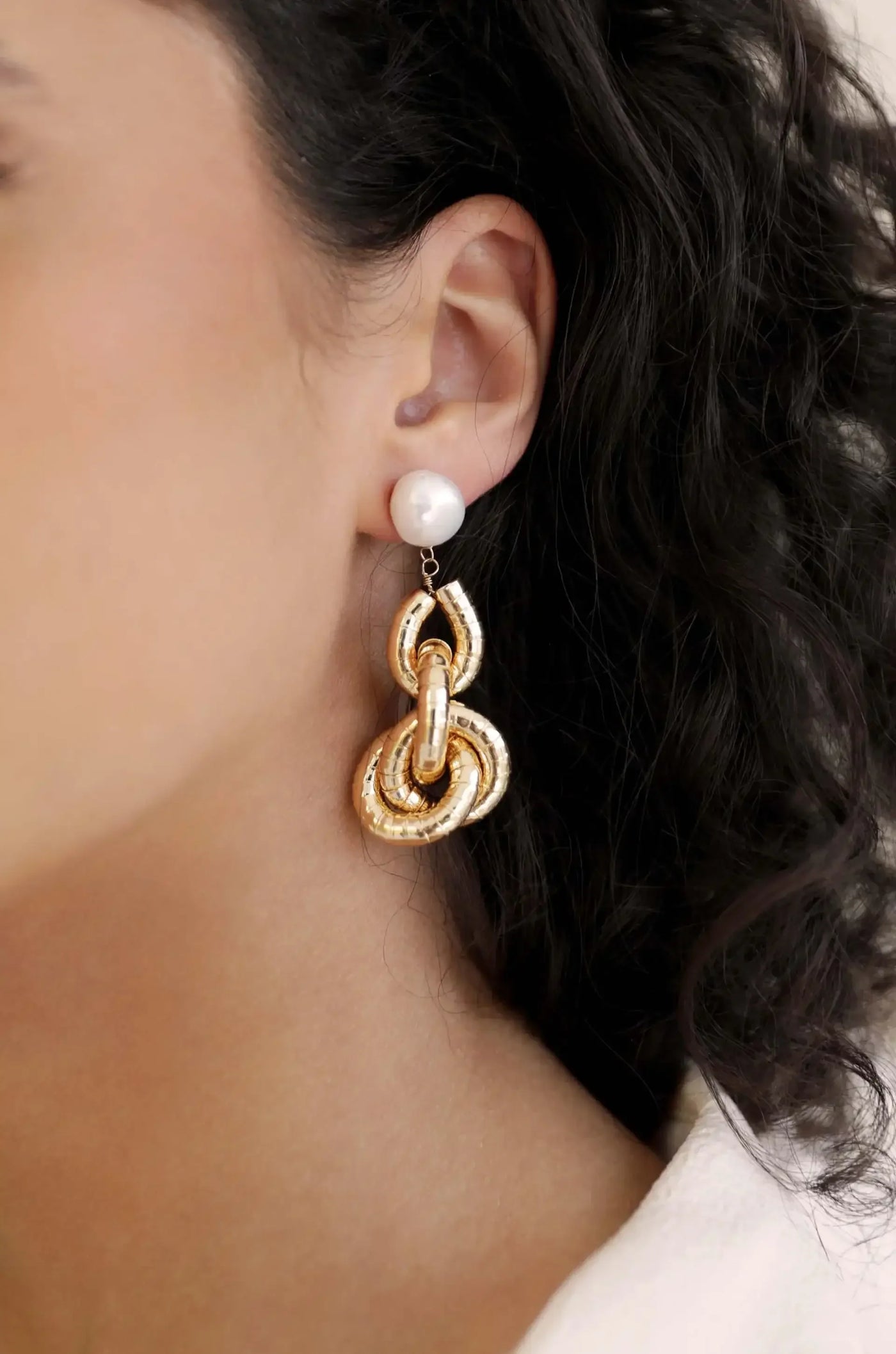 Gold and pearl earrings dangle elegantly from a woman's ear, highlighting intricate designs while complementing her curly hair and minimalist outfit in a softly lit environment.