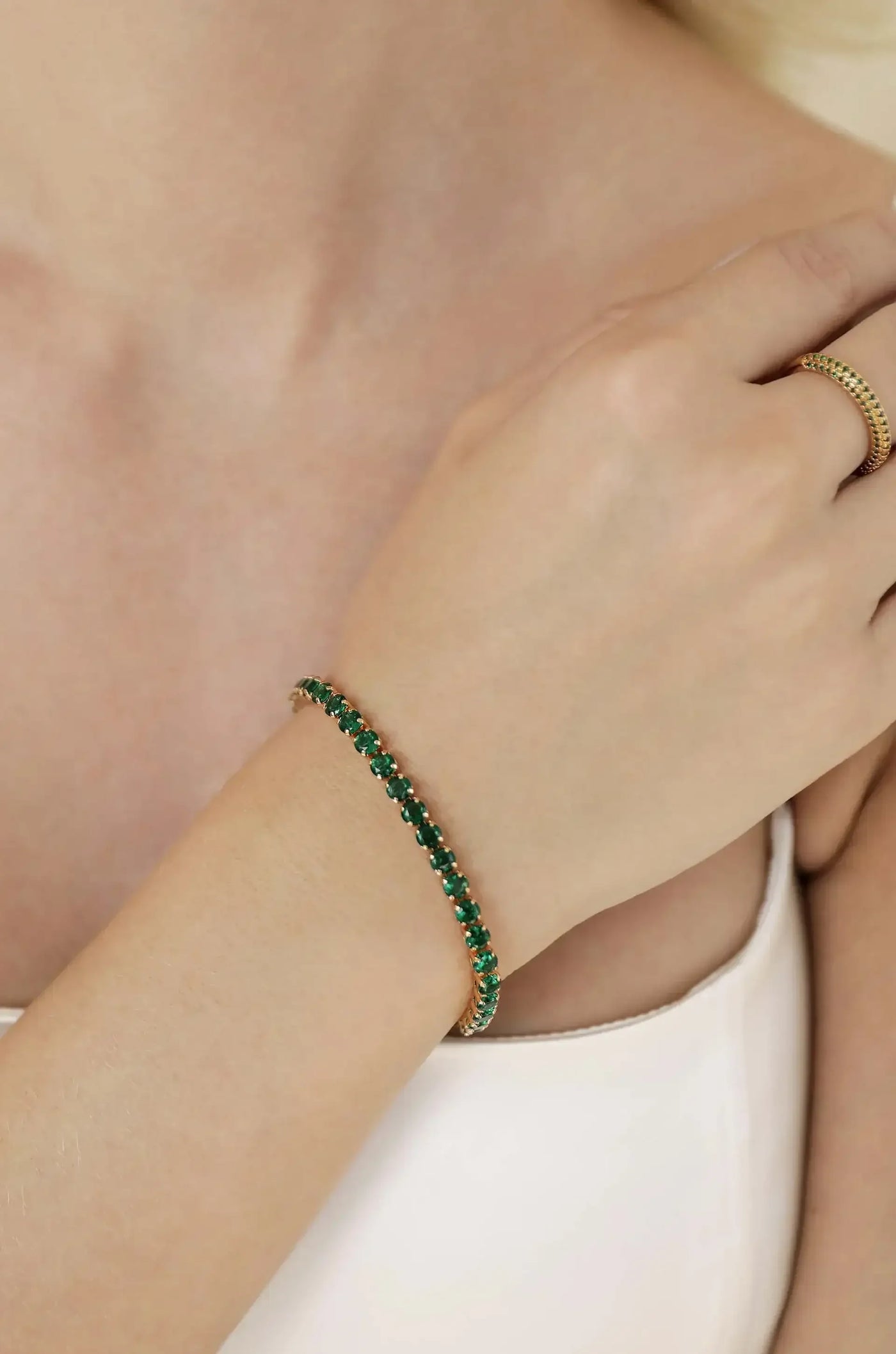 A delicate gold bracelet set with green gemstones adorns a woman's wrist. She gently touches her arm, standing against a soft, neutral background that enhances the jewelry's elegance.