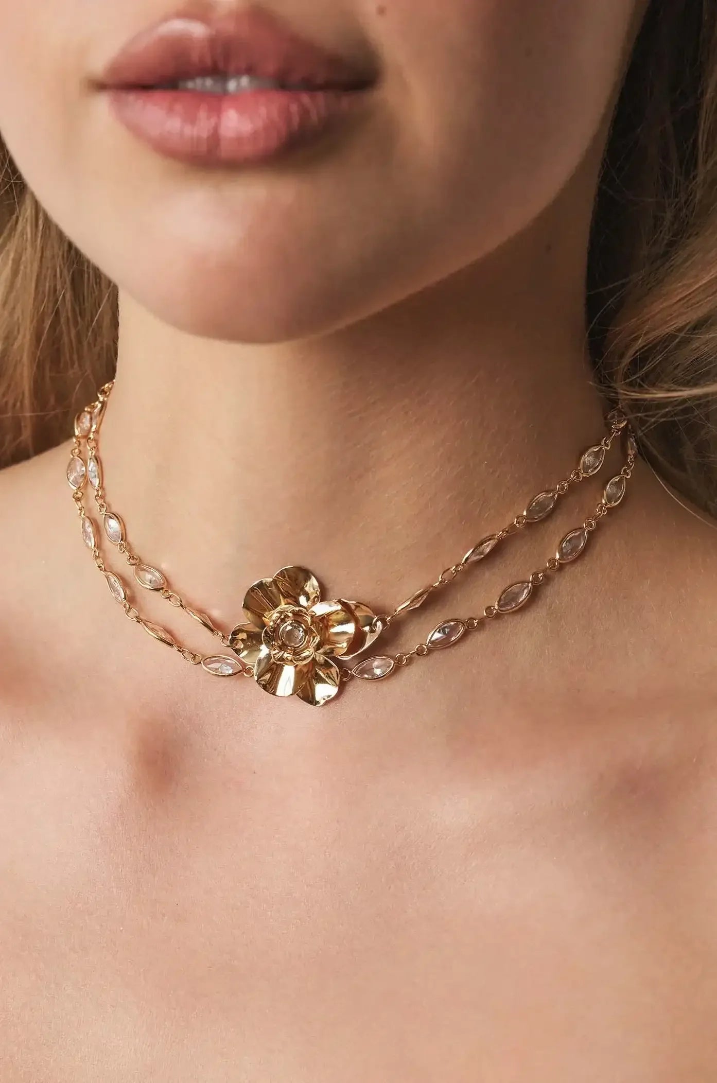 A golden flower necklace is elegantly displayed around a woman's neck, accentuating her collarbone, while she looks toward the camera with slightly parted lips, set against a soft, neutral background.