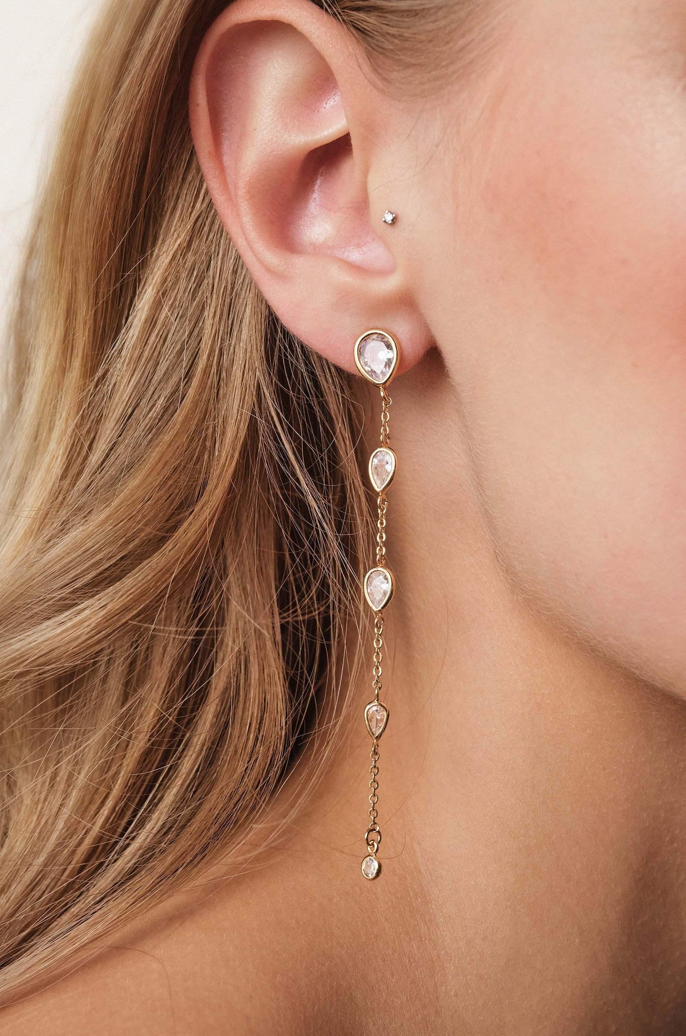 A close-up of a woman's ear showcases a delicate dangling earring with multiple teardrop-shaped gems, set in gold, complementing her long, wavy hair and skin tone.