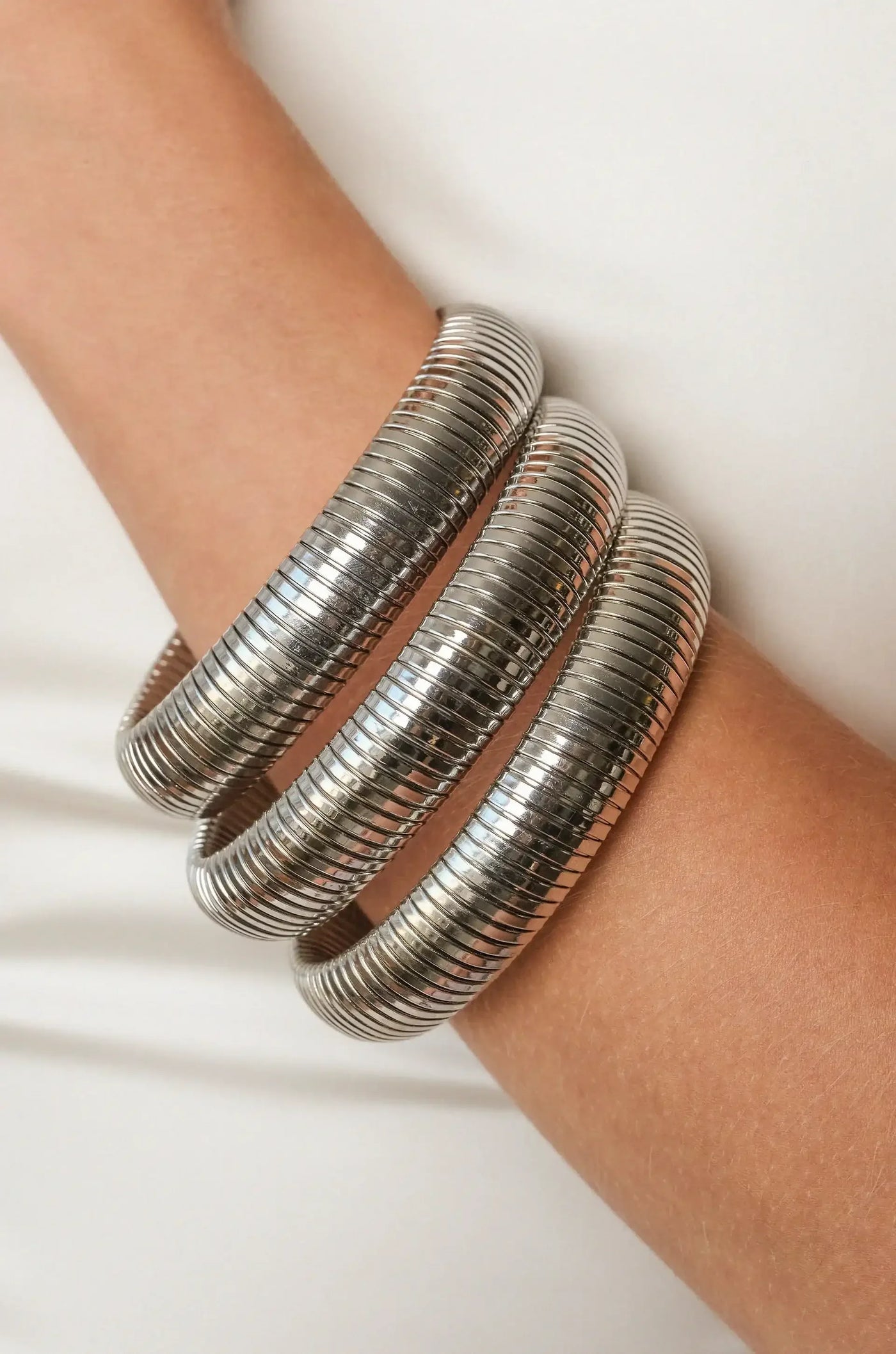 A metallic bangle, consisting of three stacked, ribbed bands, adorns a light-skinned arm. The background is neutral, emphasizing the shiny texture of the jewelry.