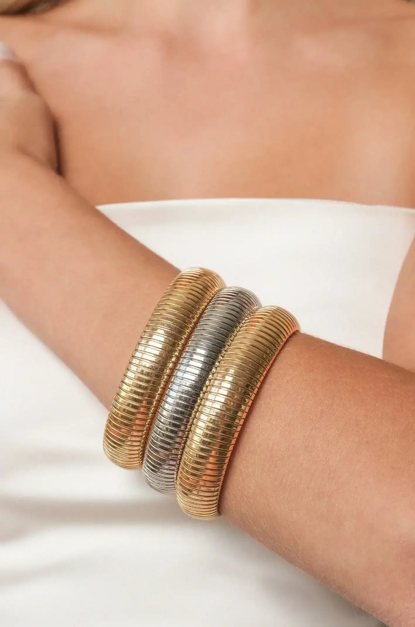 Three metallic banglesâ€”two gold and one silverâ€”are stacked on a bare arm. The background features a soft, neutral tone, emphasizing the jewelry's shine and texture.