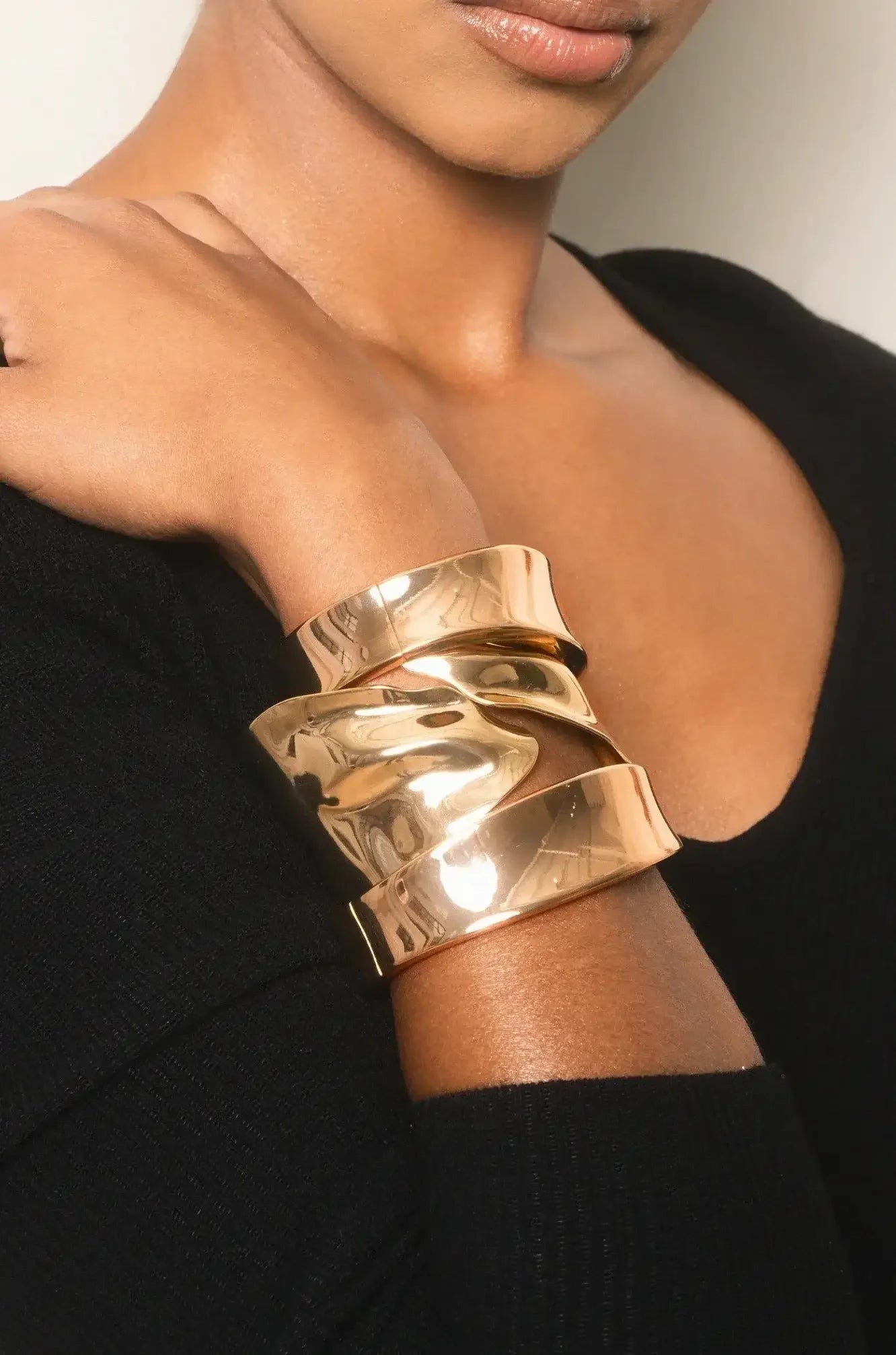 A gold-toned cuff bracelet adorned with wavy layers is worn on a person's wrist. The individual is partially visible, wearing a black sweater. The background appears neutral and unobtrusive.