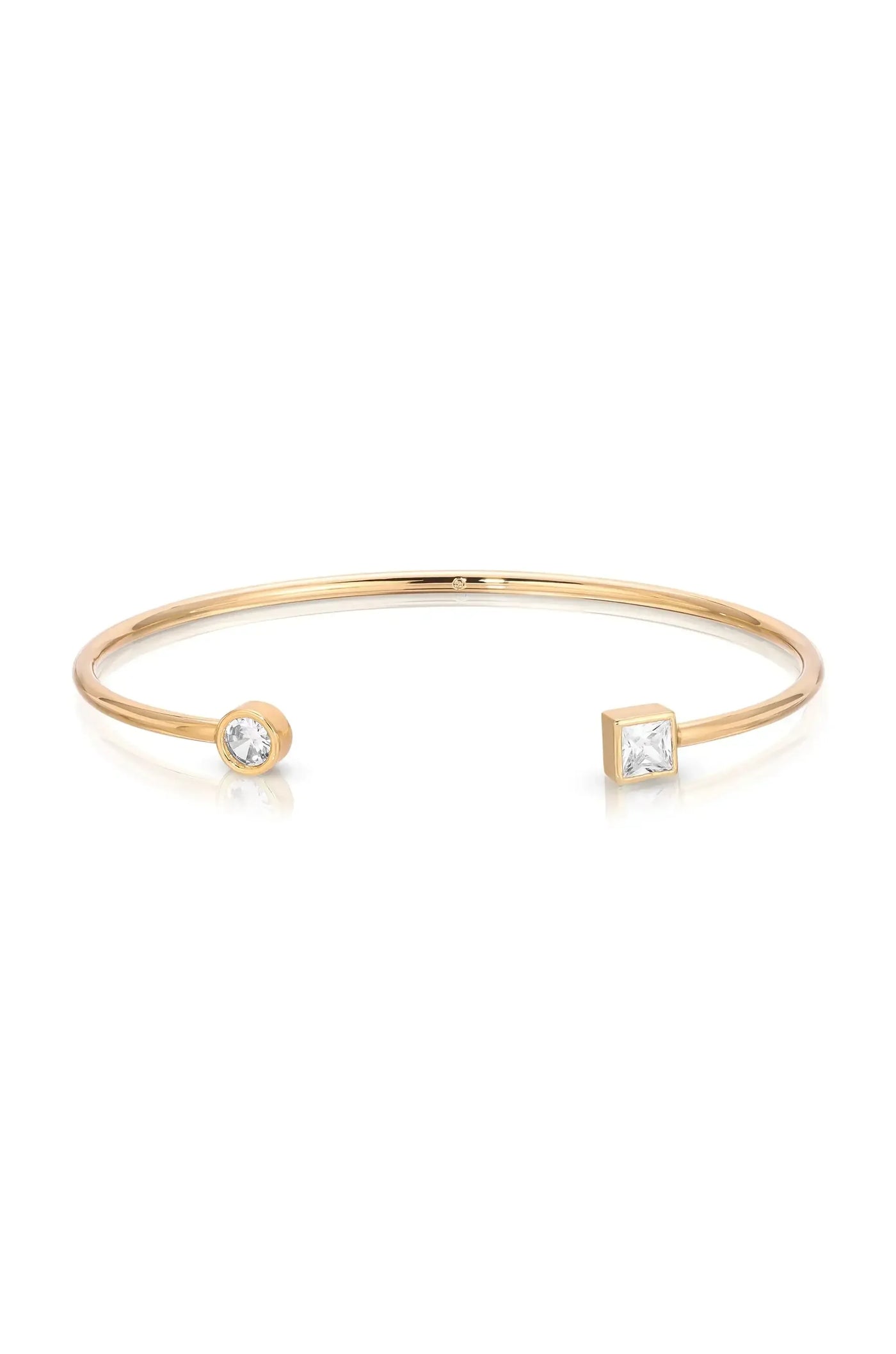A gold cuff bracelet features two decorative ends, one round and one square, each set with a gemstone. The bracelet has a sleek, minimalist design, shining against a white background.