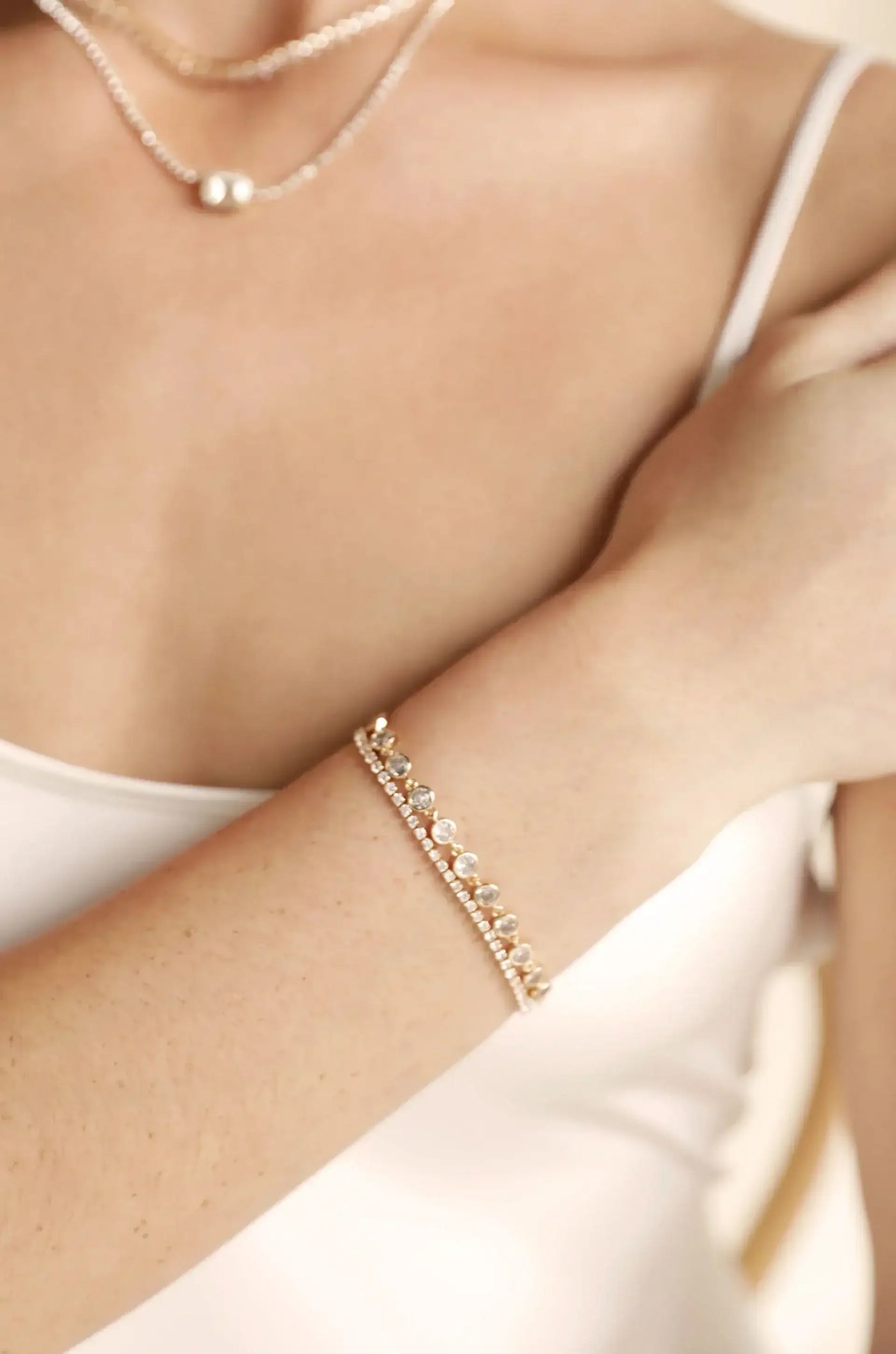A delicate gold bracelet with sparkling stones adorns a wrist. The wearer gently rests their hand against their shoulder, showcasing the elegance of the jewelry against a softly blurred background.