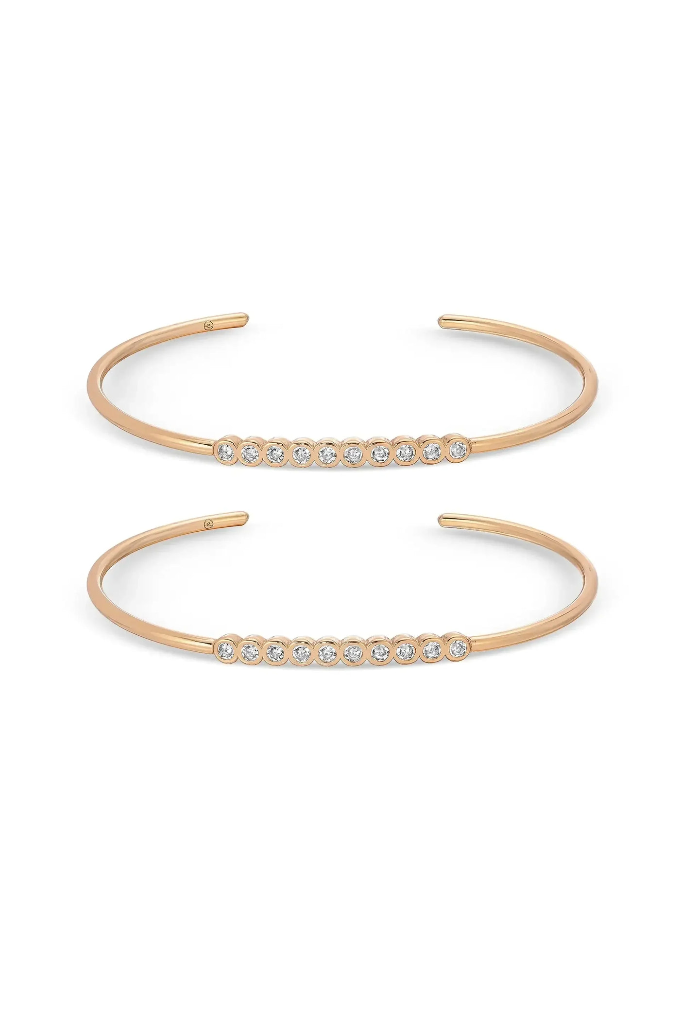 Two thin, open-ended bracelets are displayed side by side. The upper bracelet features a row of small, sparkling stones, while the lower one showcases a similar design with more stones, both set in a gold-tone metal.