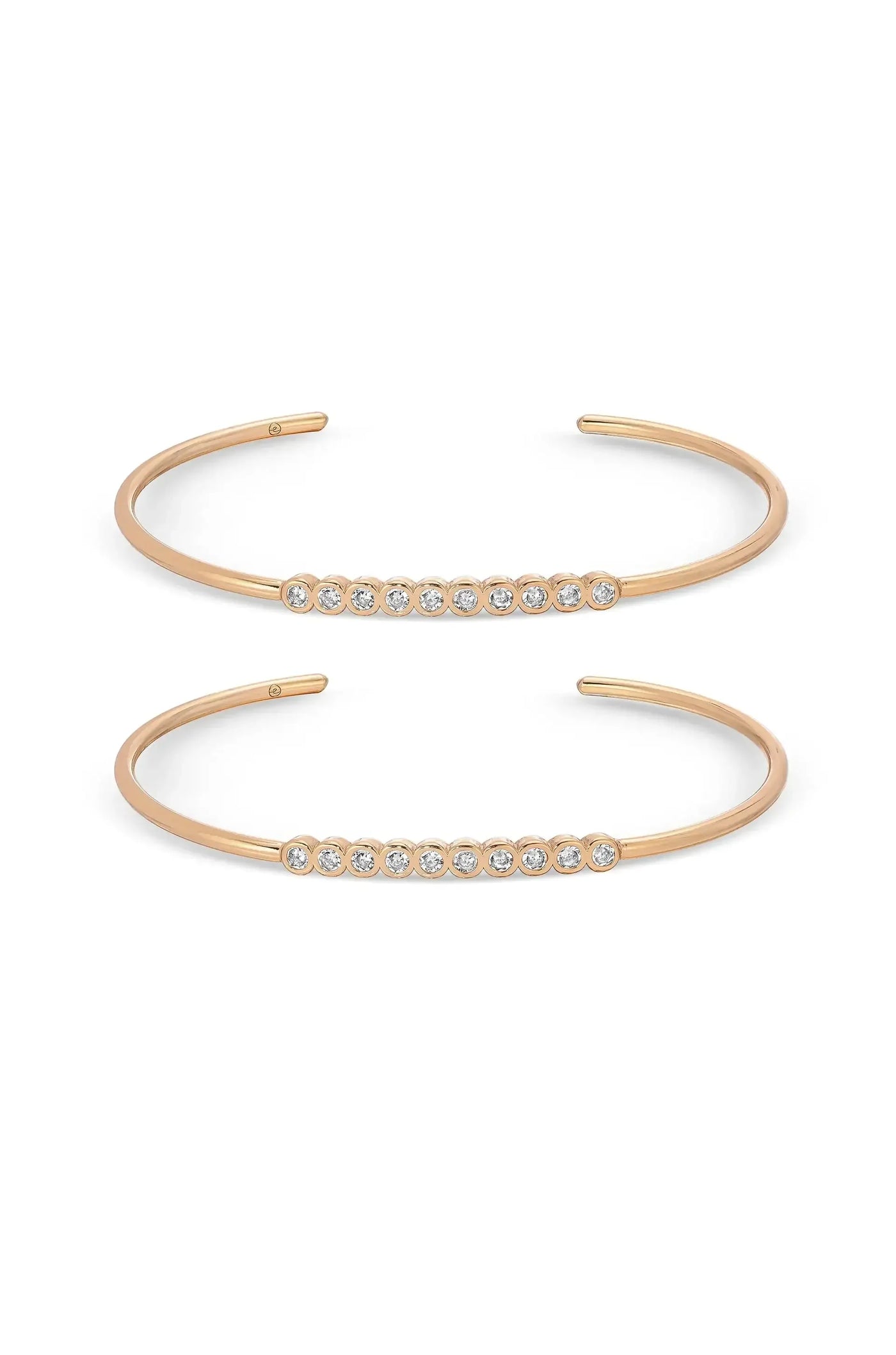 Two thin, open-ended bracelets are displayed side by side. The upper bracelet features a row of small, sparkling stones, while the lower one showcases a similar design with more stones, both set in a gold-tone metal.