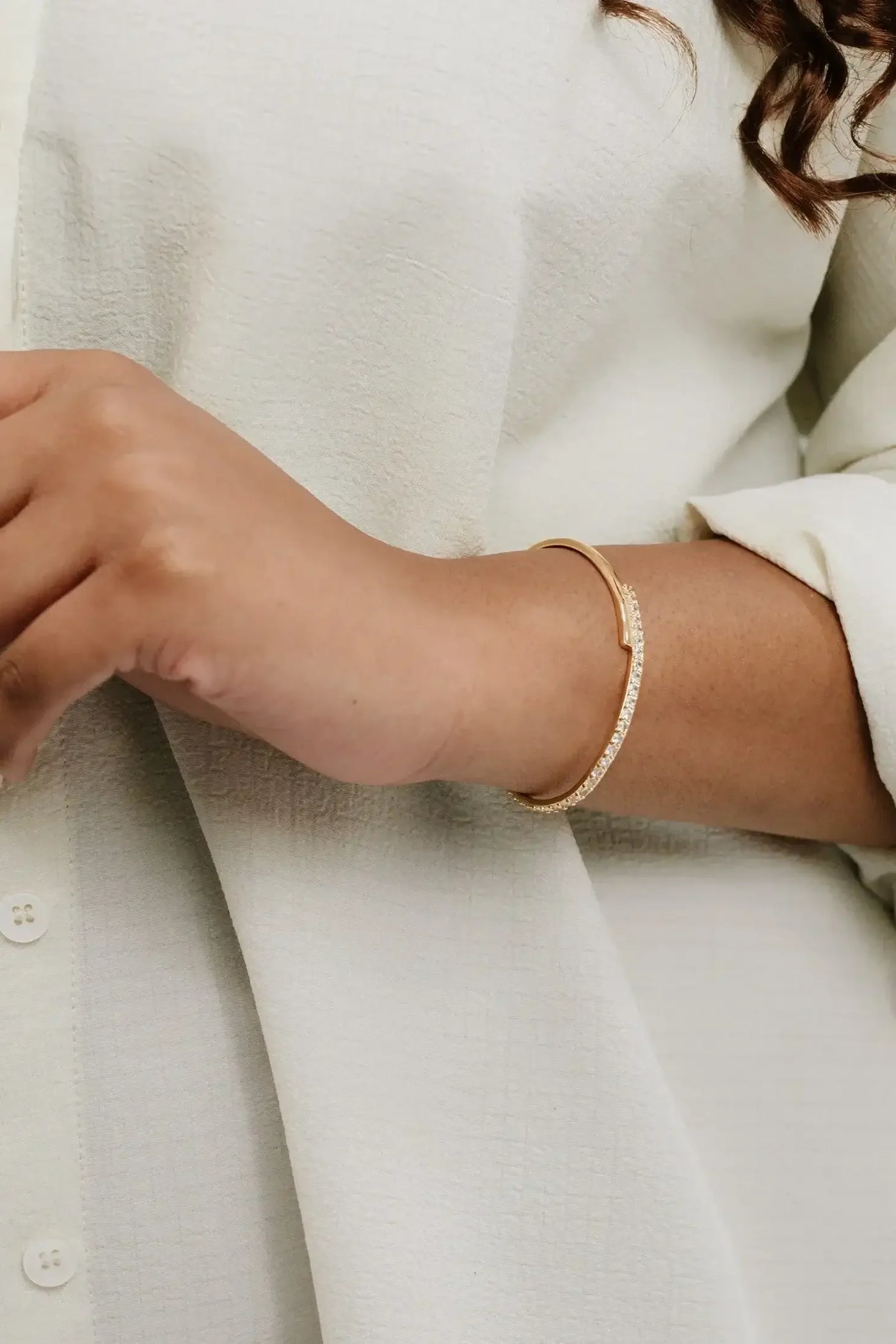 A hand is resting on a light-colored shirt, showcasing a gold bracelet adorned with sparkling stones. The shirt has a textured pattern, providing a soft, elegant backdrop.