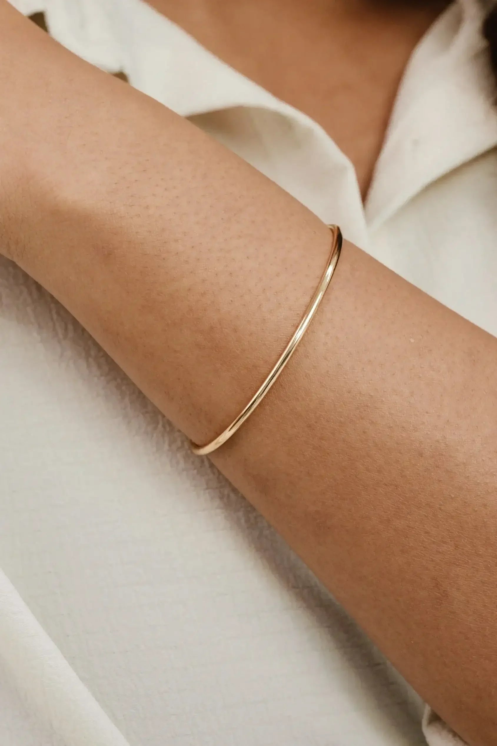 A slender gold bangle adorns a smooth, tanned arm. The arm is relaxed against a textured, light-colored surface, enhancing the bangle's elegant simplicity and shine.