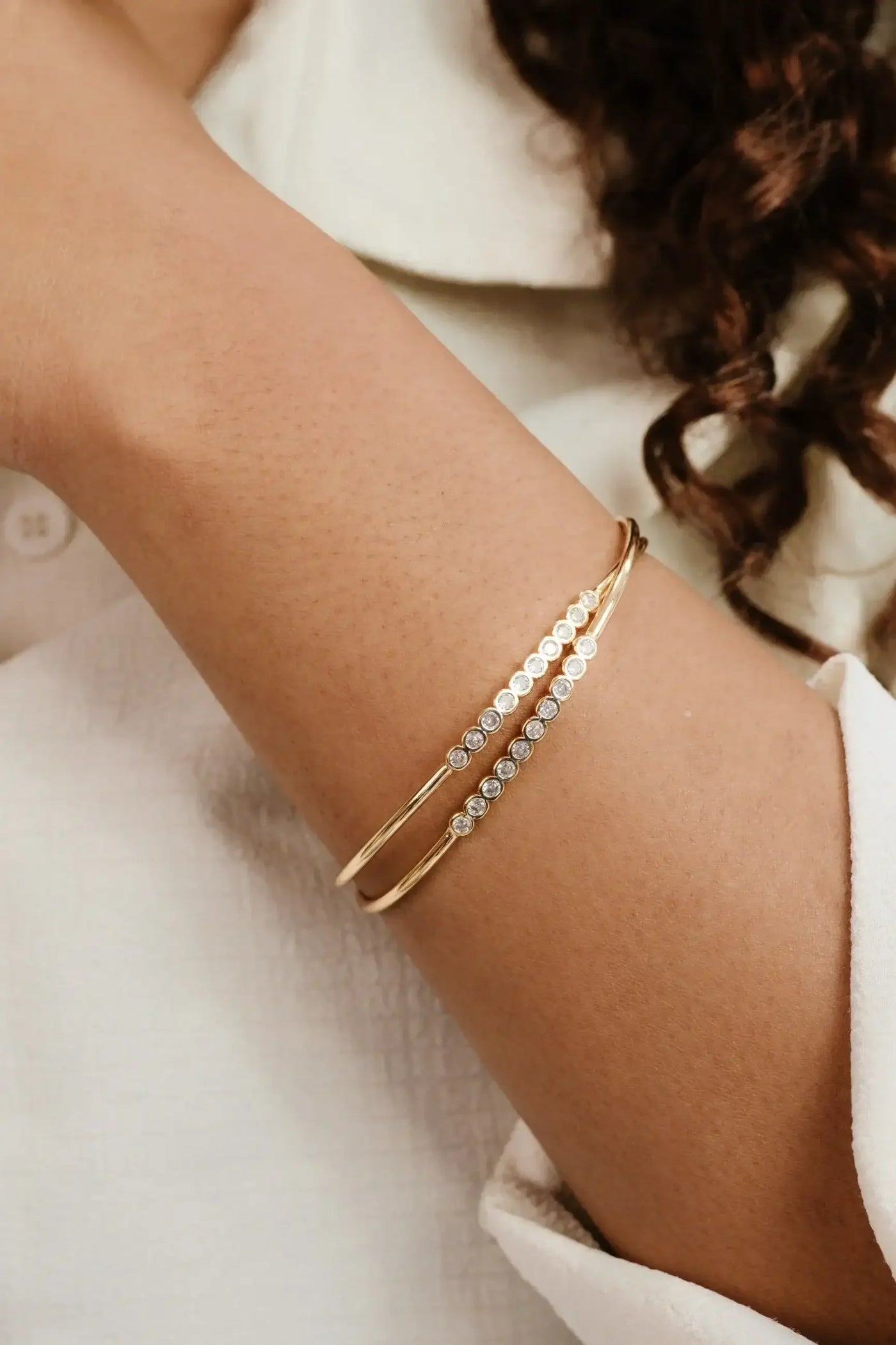 A gold bangle with embedded clear stones adorns a wrist. The arm rests against a textured white surface, and a light-colored garment partially covers the wrist.