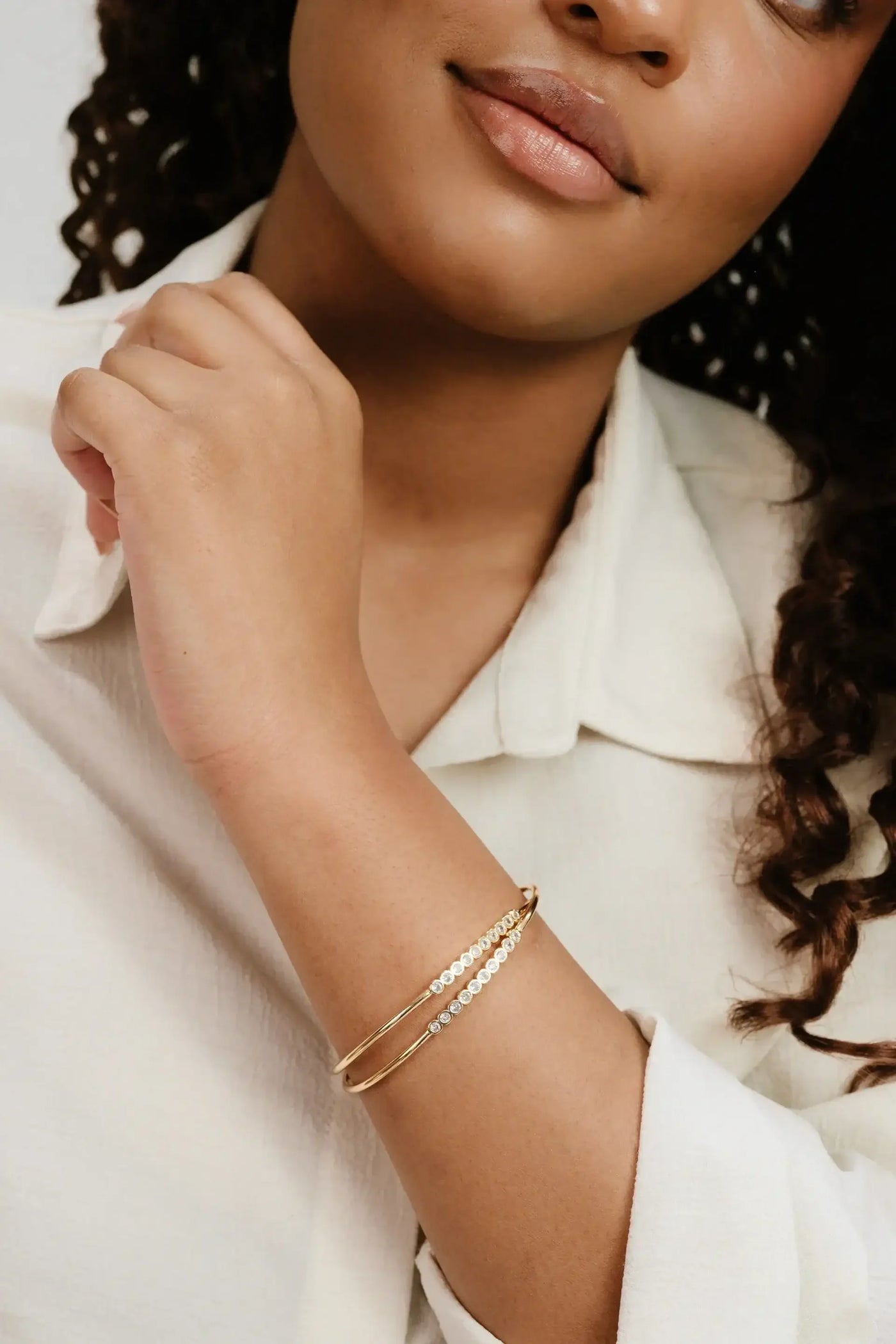 A woman's hand gently rests against her collarbone, showcasing delicate gold bracelets with sparkling details. She wears a light-colored shirt, and the background is softly muted.